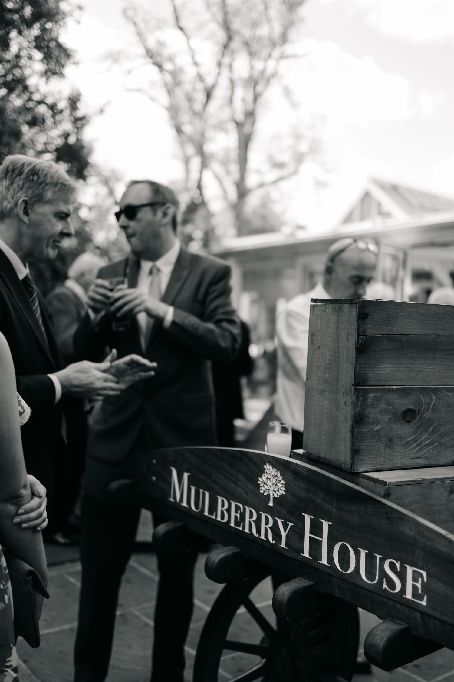Black and white photo of wooden Mulberry House venue sign with guests in background.