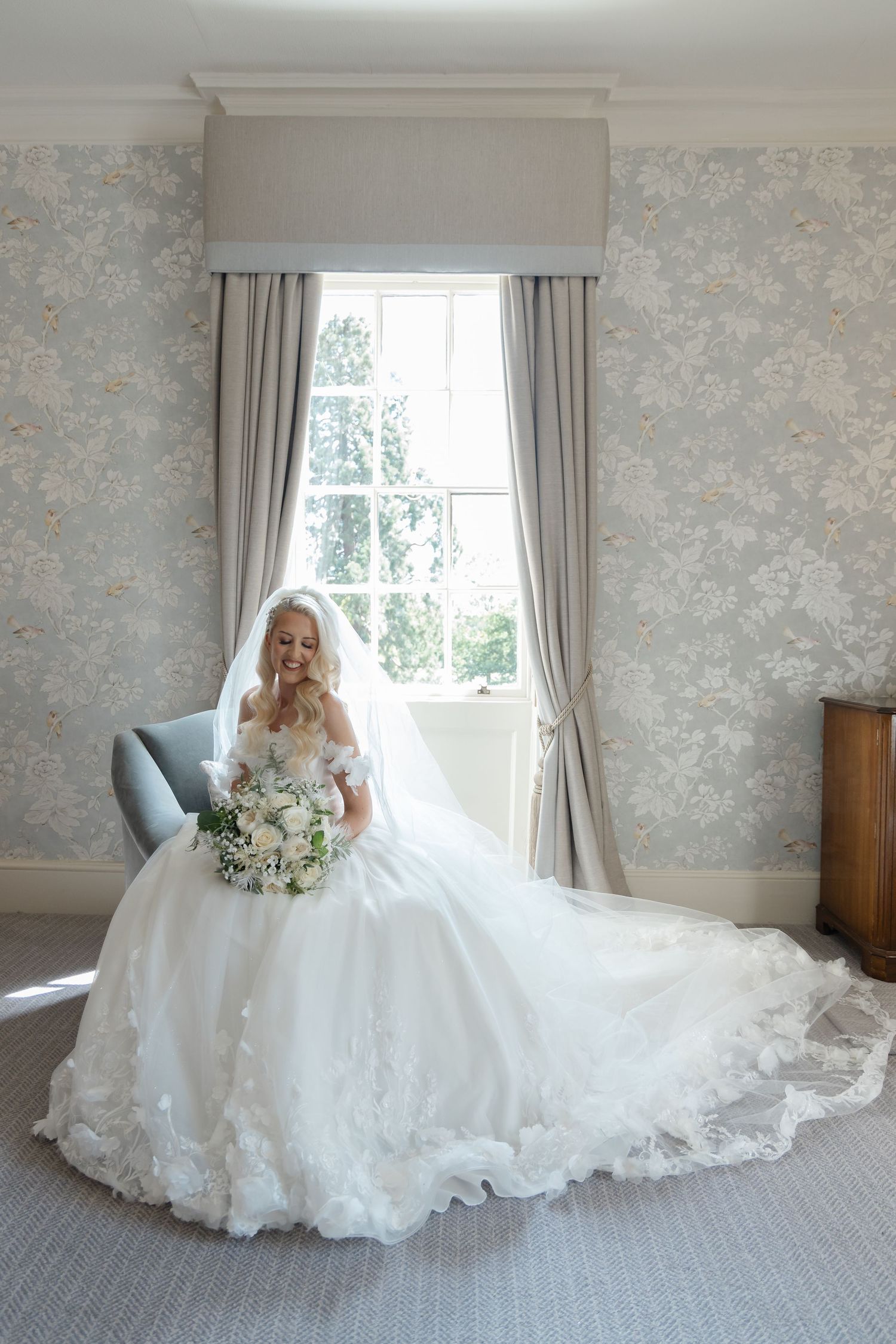 Bride in elaborate lace wedding gown poses in elegant room with floral wallpaper.