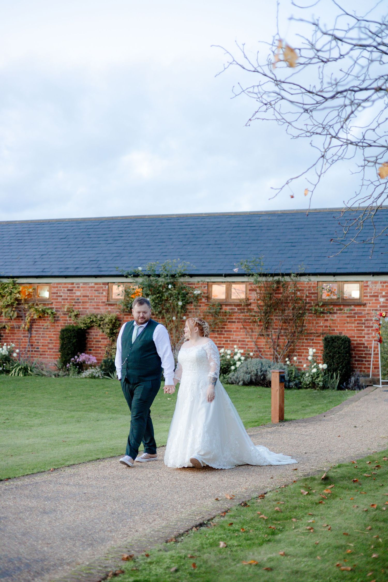 Wedding couple walks along path by brick building during outdoor photo session.