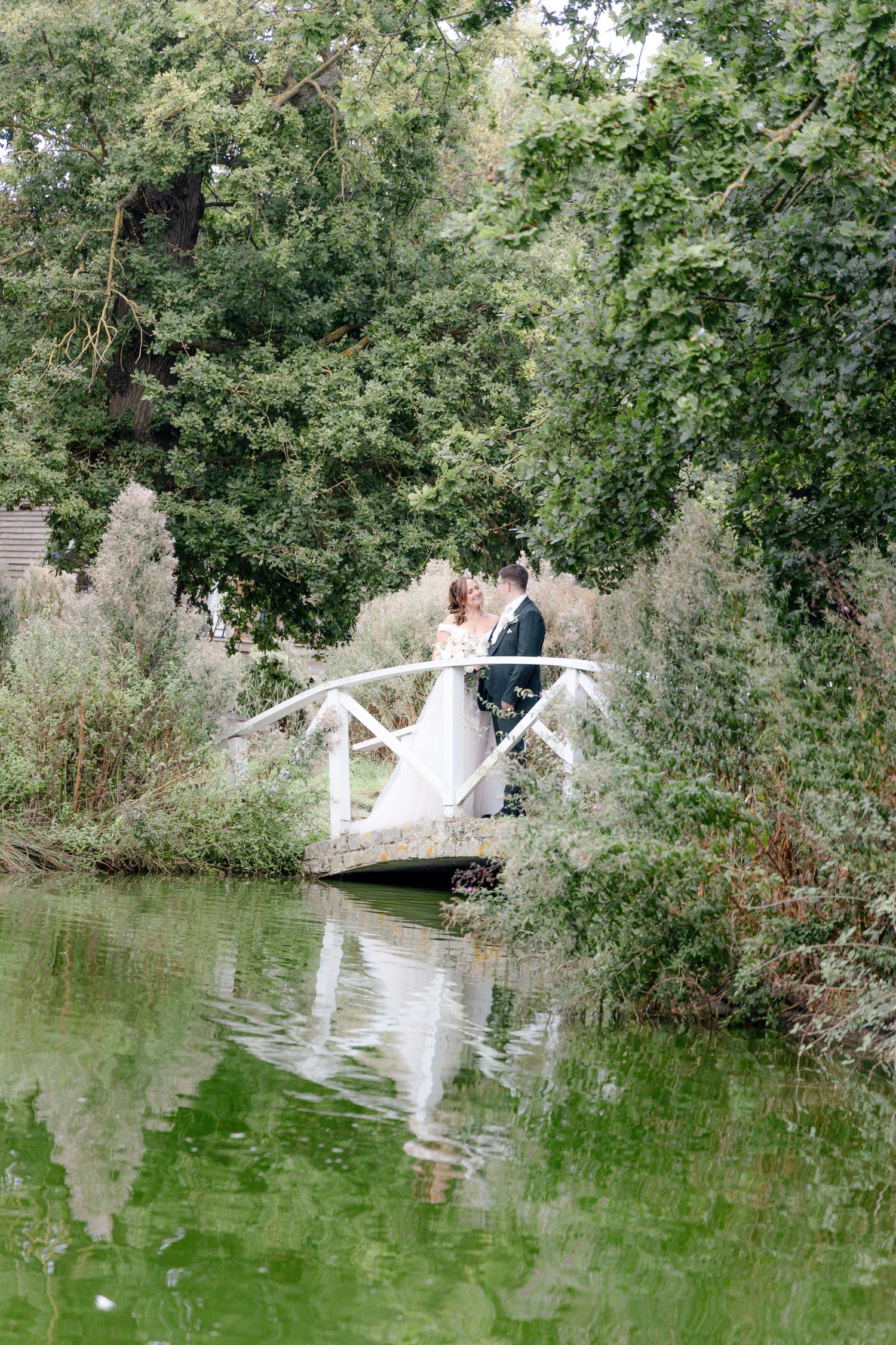 Scenic wedding photo shoot by a tranquil pond with white bridge and lush greenery.