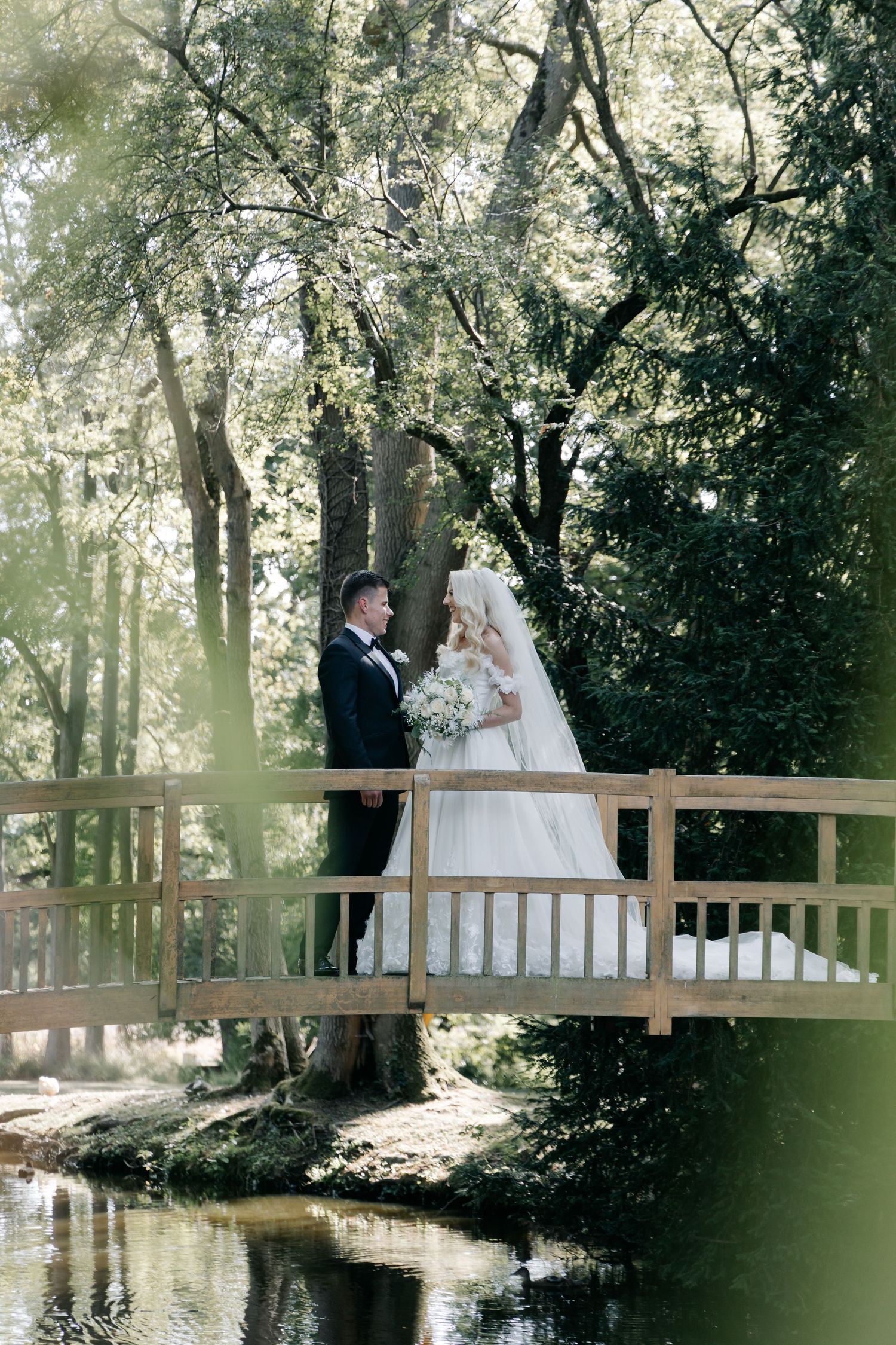 Wedding scene on wooden bridge over serene pond surrounded by trees.