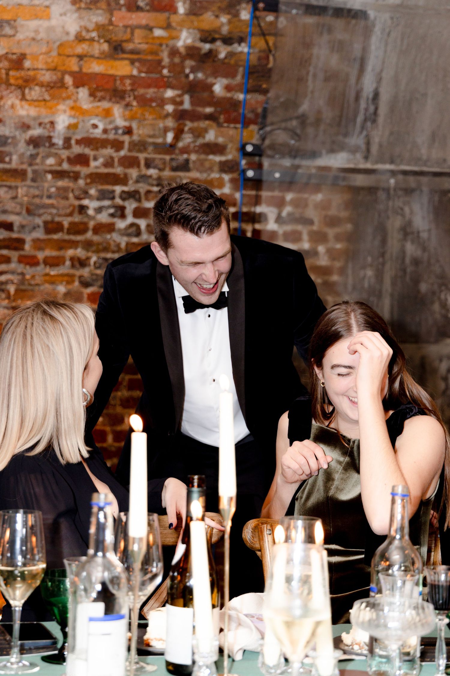 Formal dinner party guests laughing and socializing at a rustic venue with exposed brick walls.