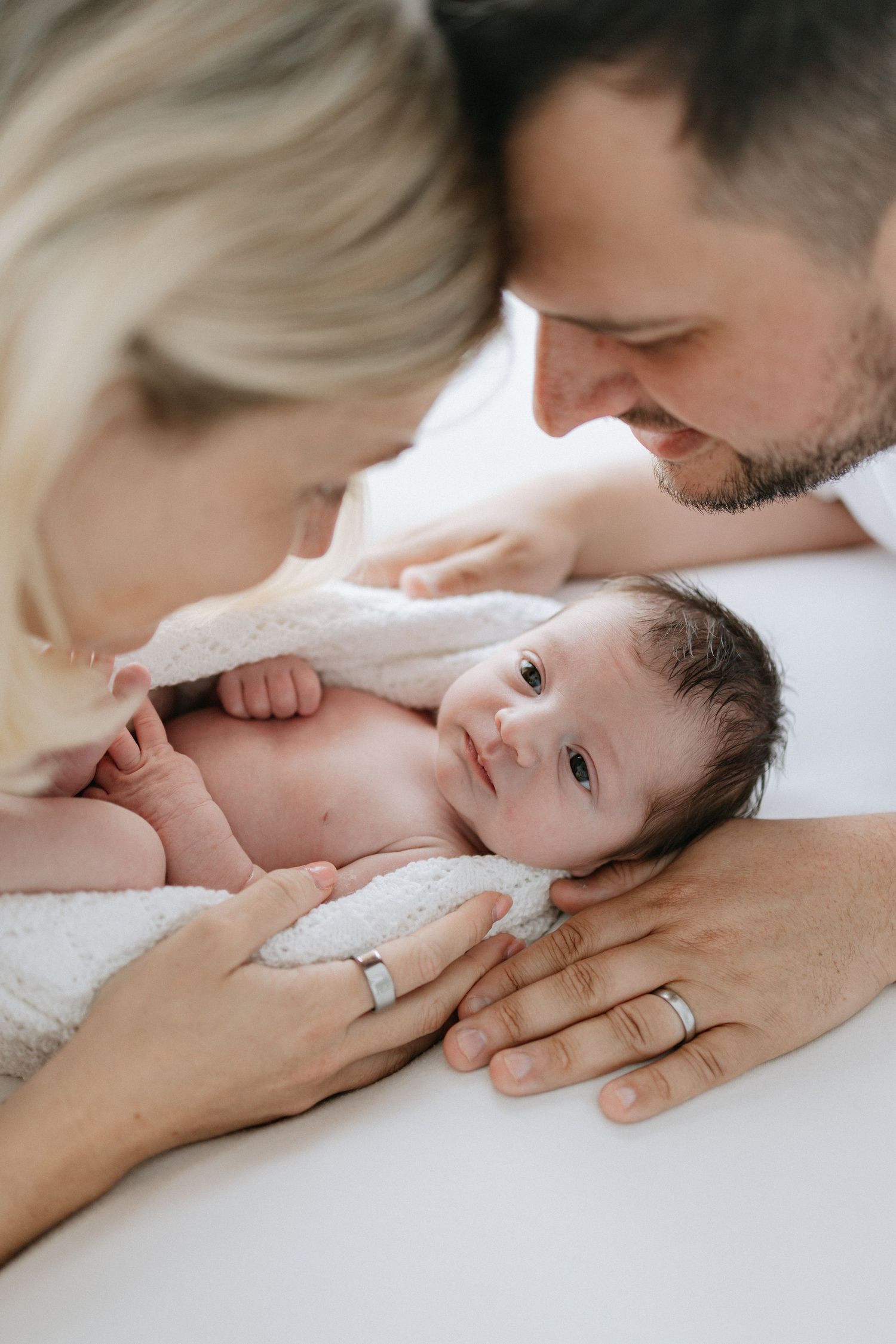 A couple with wedding rings lovingly gazes at their newborn baby lying on a white blanket.