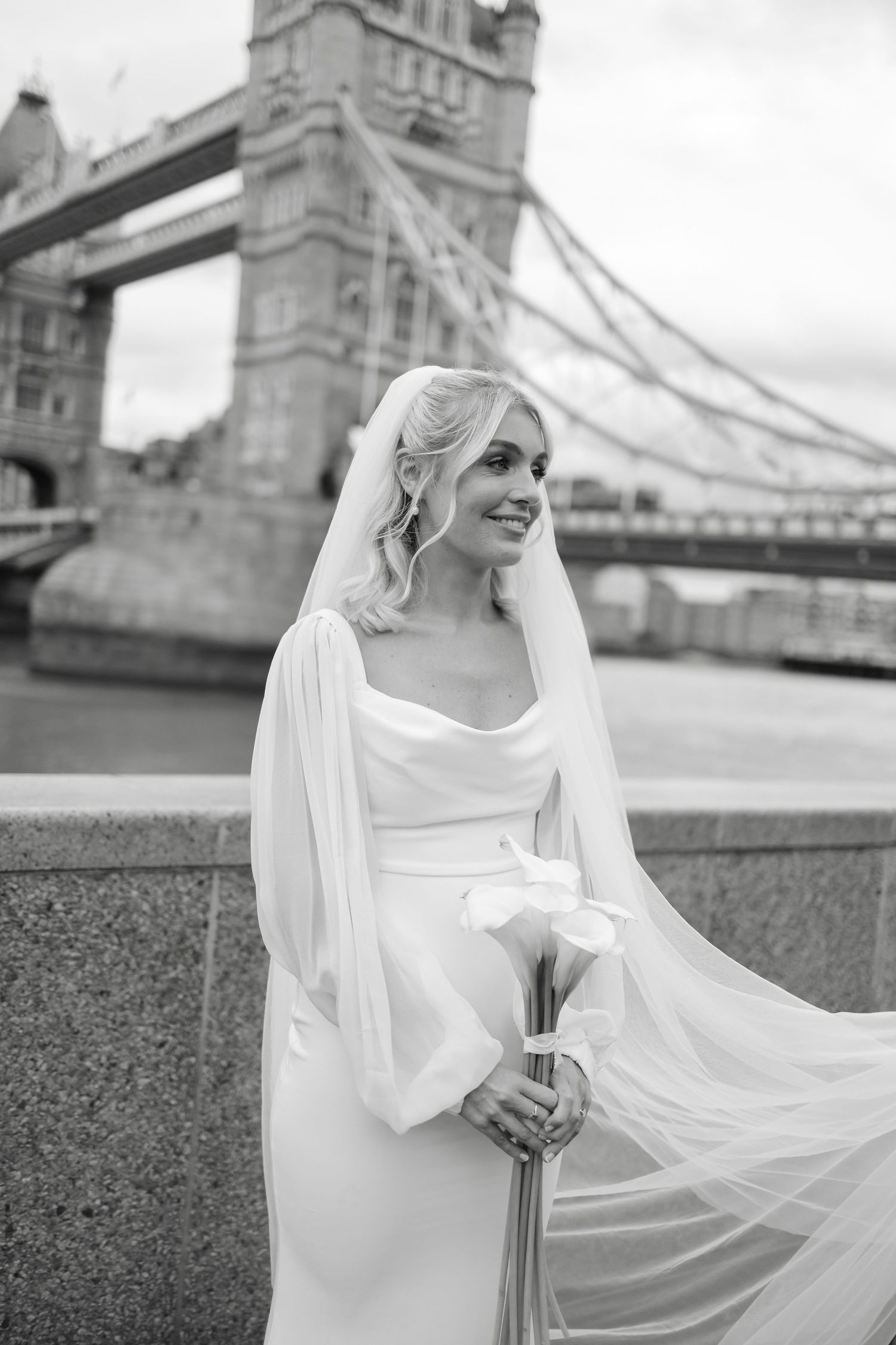 Elegant bride in white dress and flowing veil poses near London's Tower Bridge.