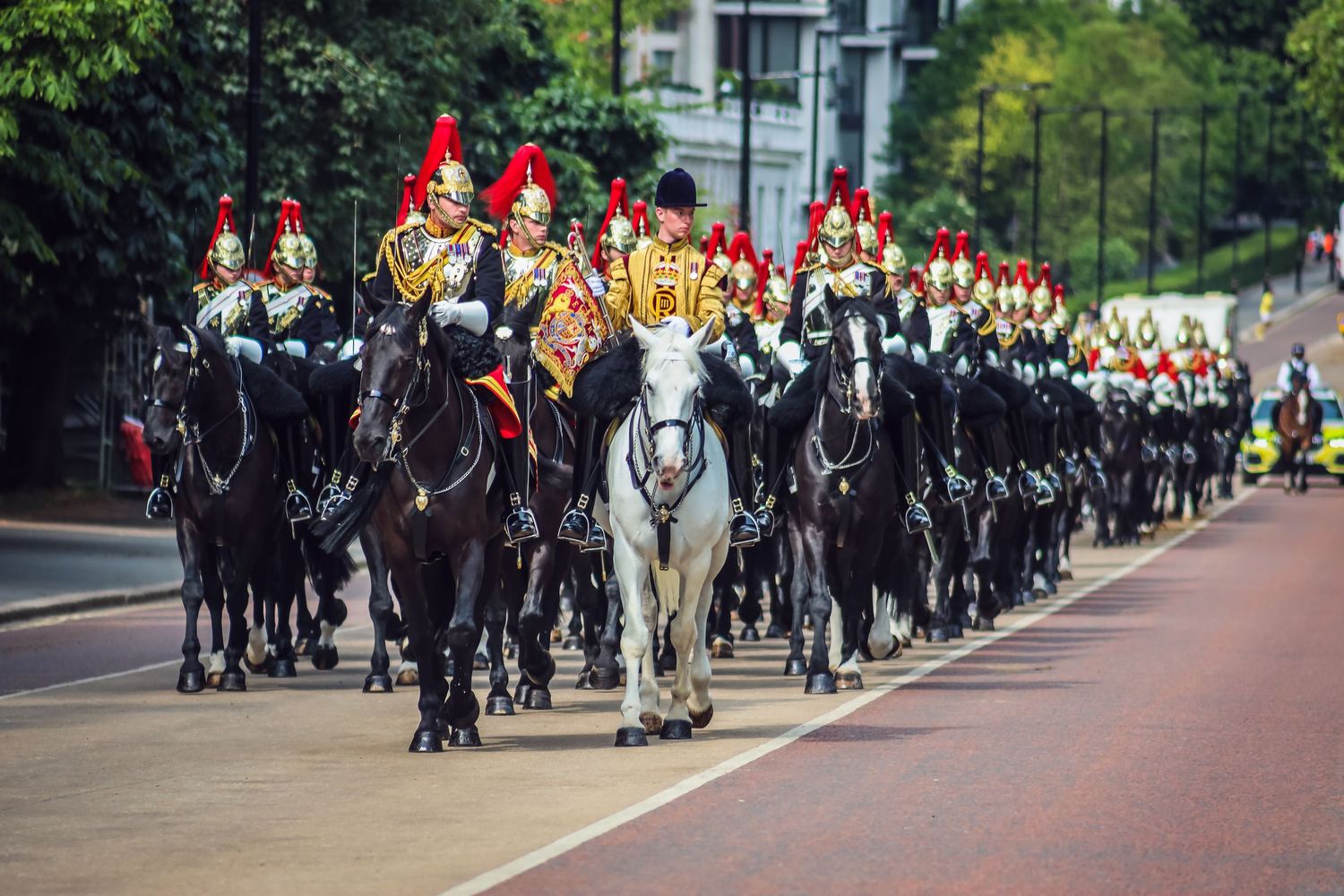 Military parade procession with mounted guards and ceremonial uniforms on a tree-lined street.