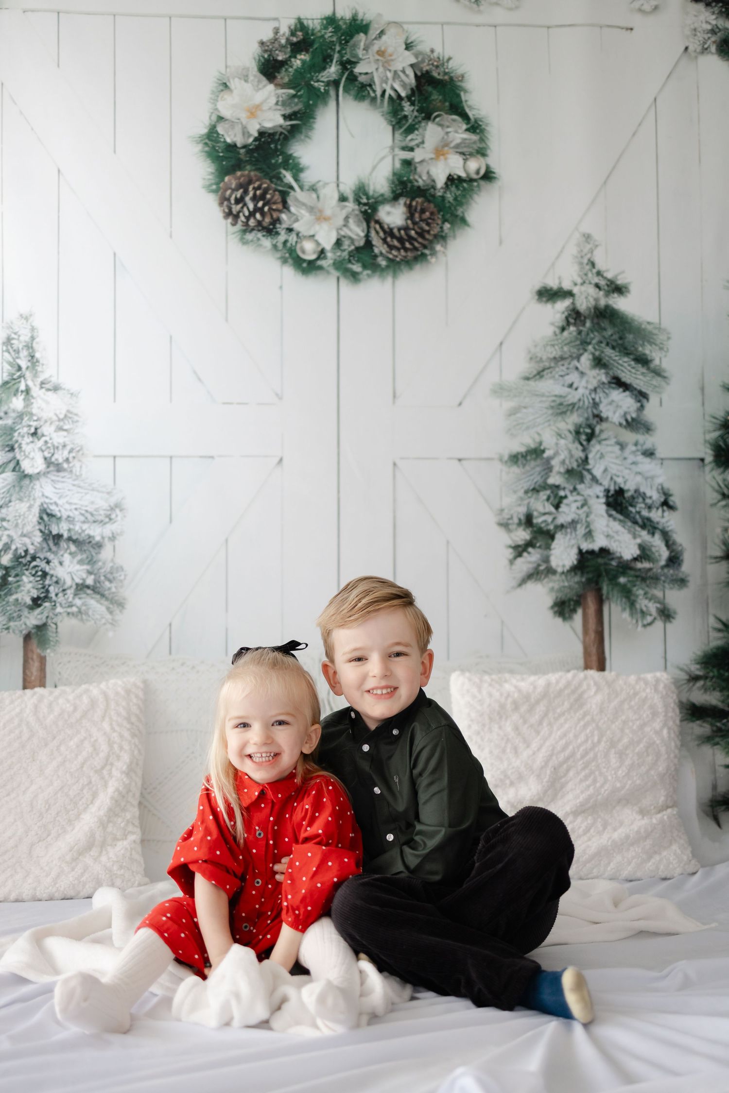 Children in festive outfits play together on white bedding with snowy evergreens in background.