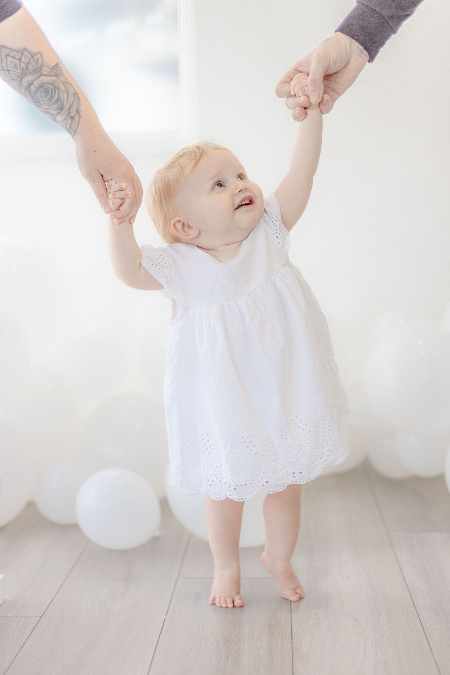 Baby in white lace dress learning to walk while holding hands with parents.