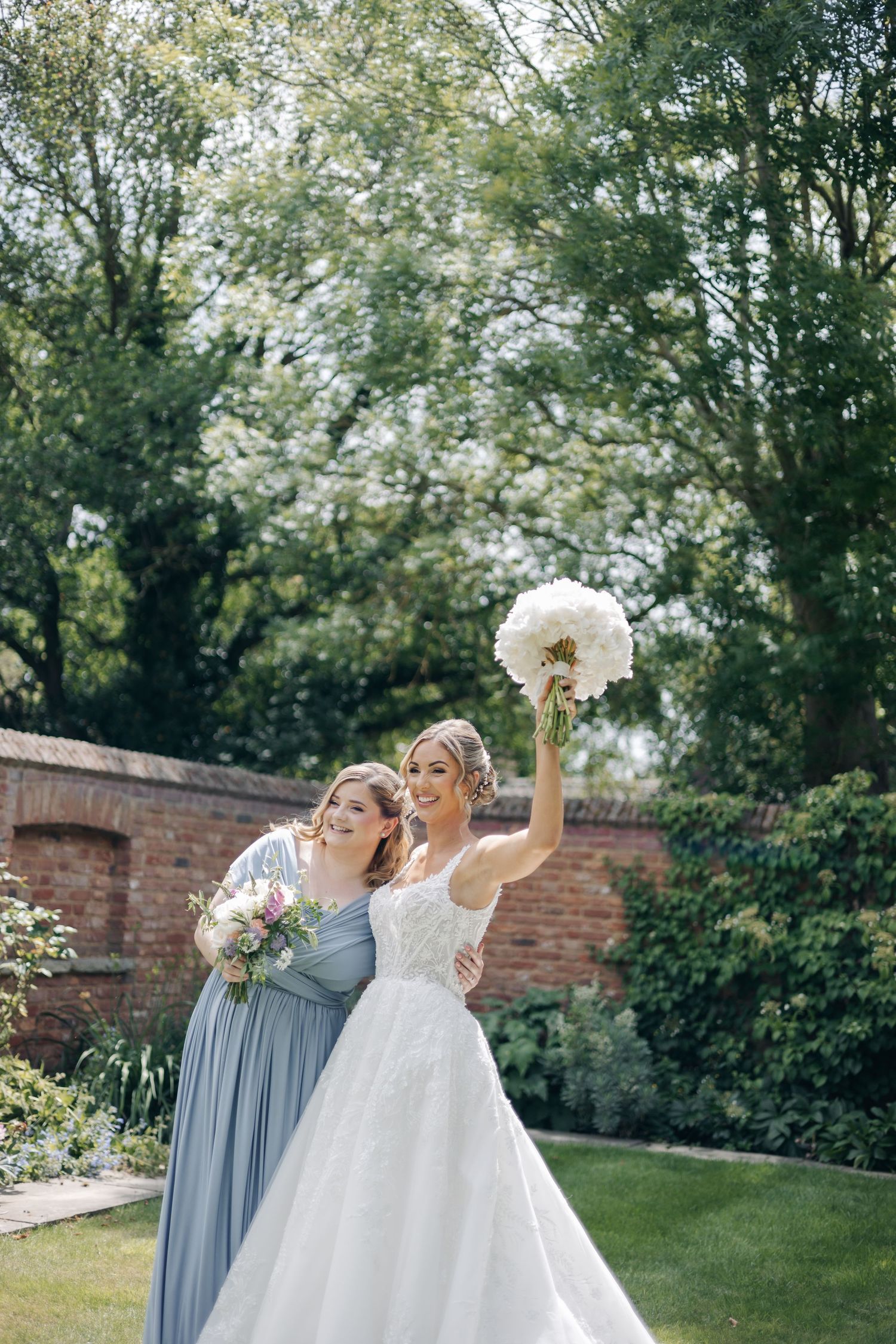 Wedding party members in gray dresses and white gown celebrate joyfully in a garden setting with brick wall backdrop.
