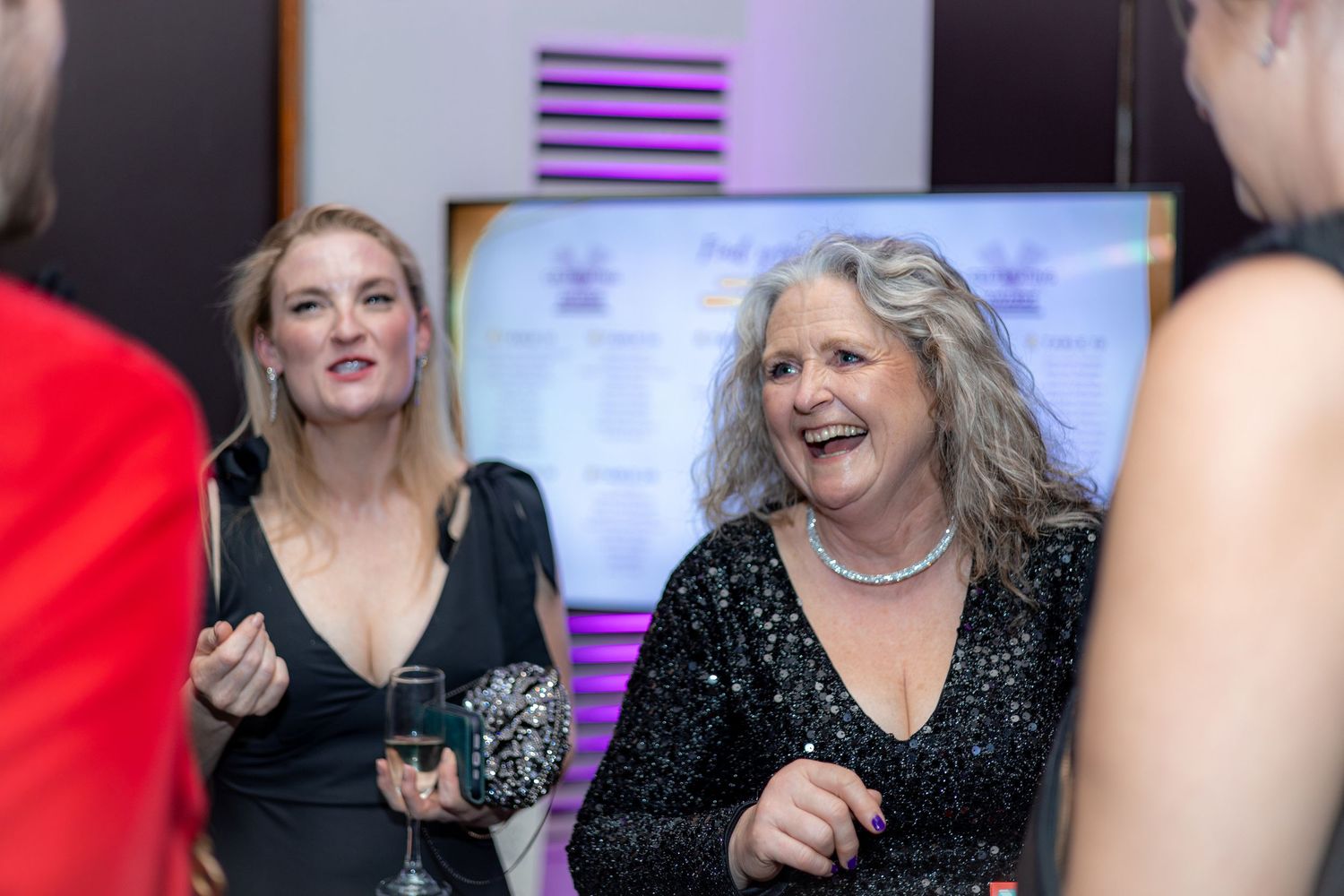 Guests in formal attire laugh and socialize at an evening event with purple decor.