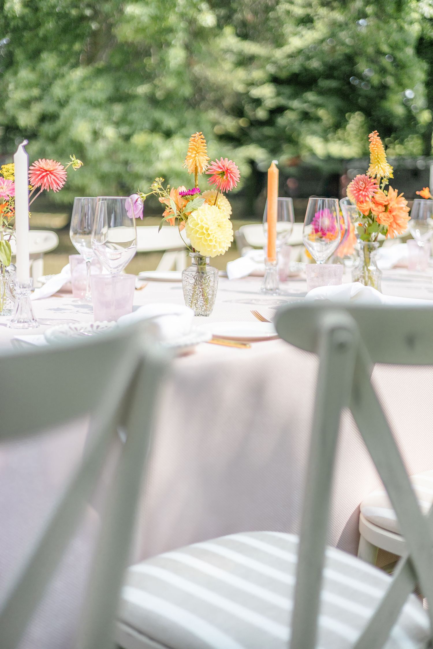 Outdoor banquet table setting with white linens, colorful flowers and orange candles in a garden.
