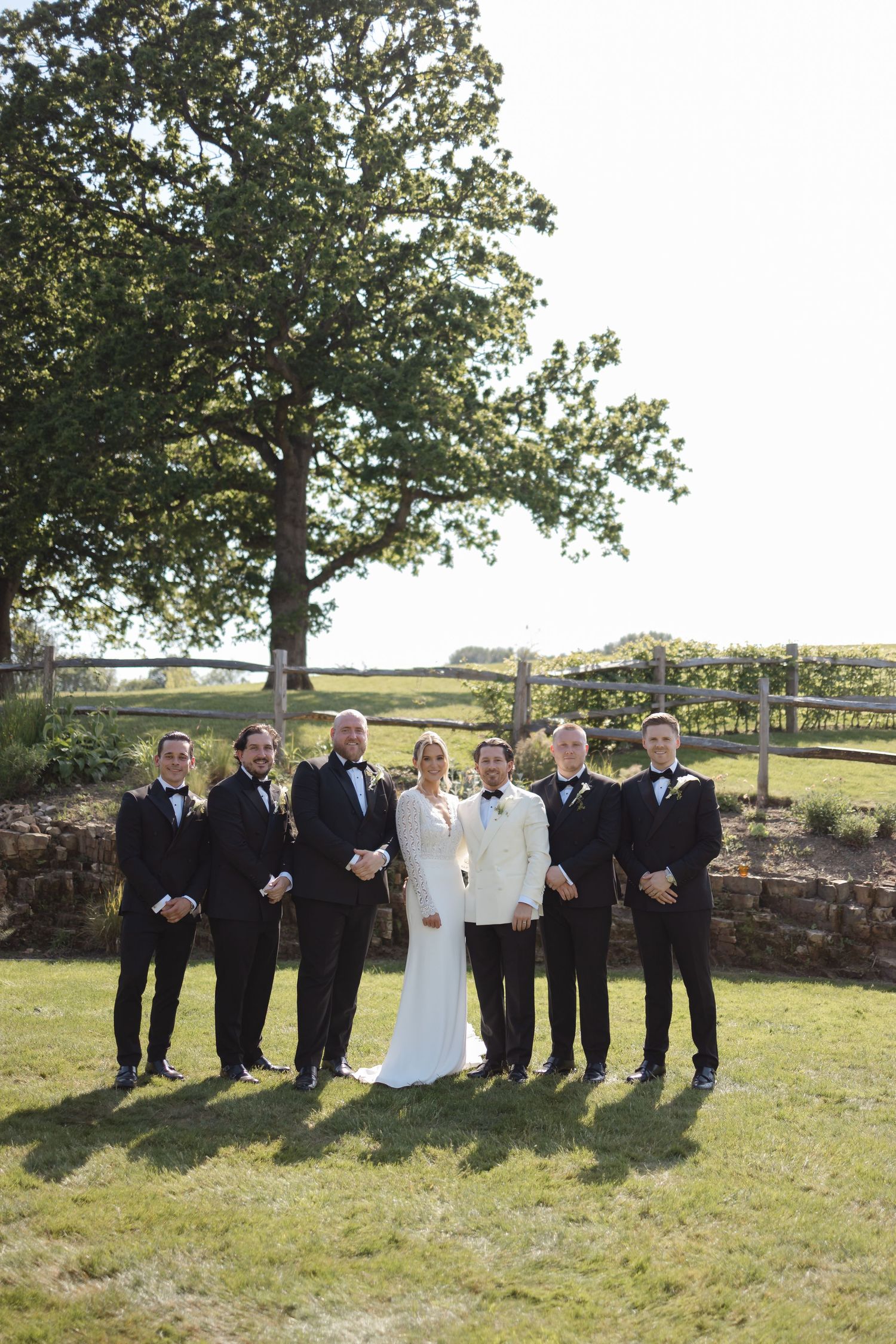 Wedding party poses outdoors with bride in white gown and groomsmen in black tuxedos beside a rustic stone wall.