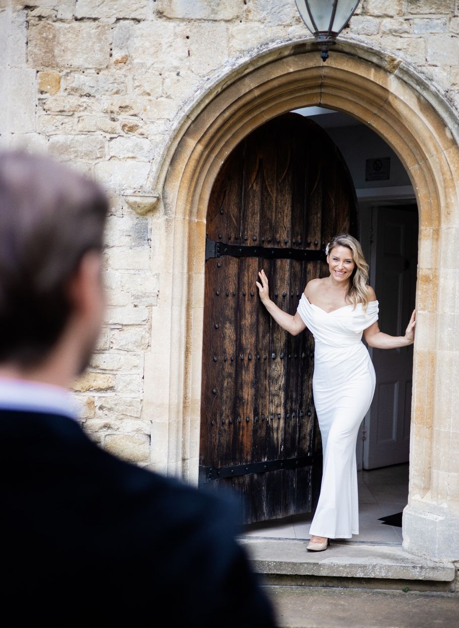Bride in white gown poses playfully in front of stone archway and wooden door.