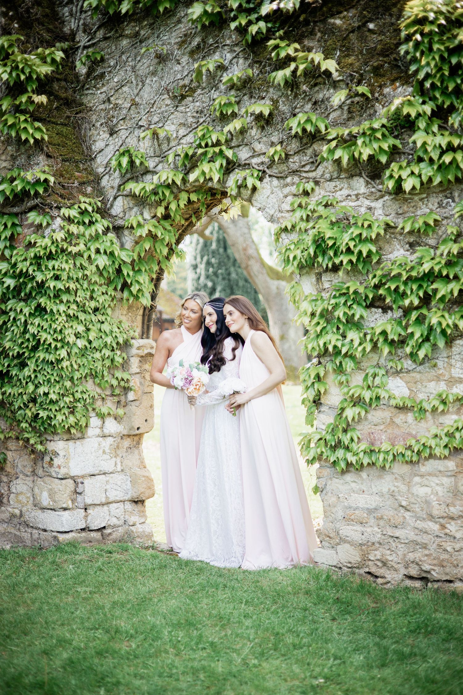 Bridesmaids in light pink and white dresses posed by vine-covered stone wall.