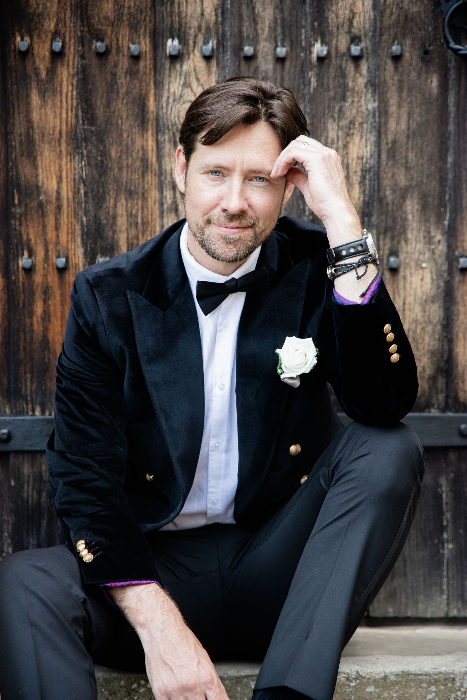 A groom in a black velvet tuxedo and bowtie poses against a rustic wooden door with decorative studs.