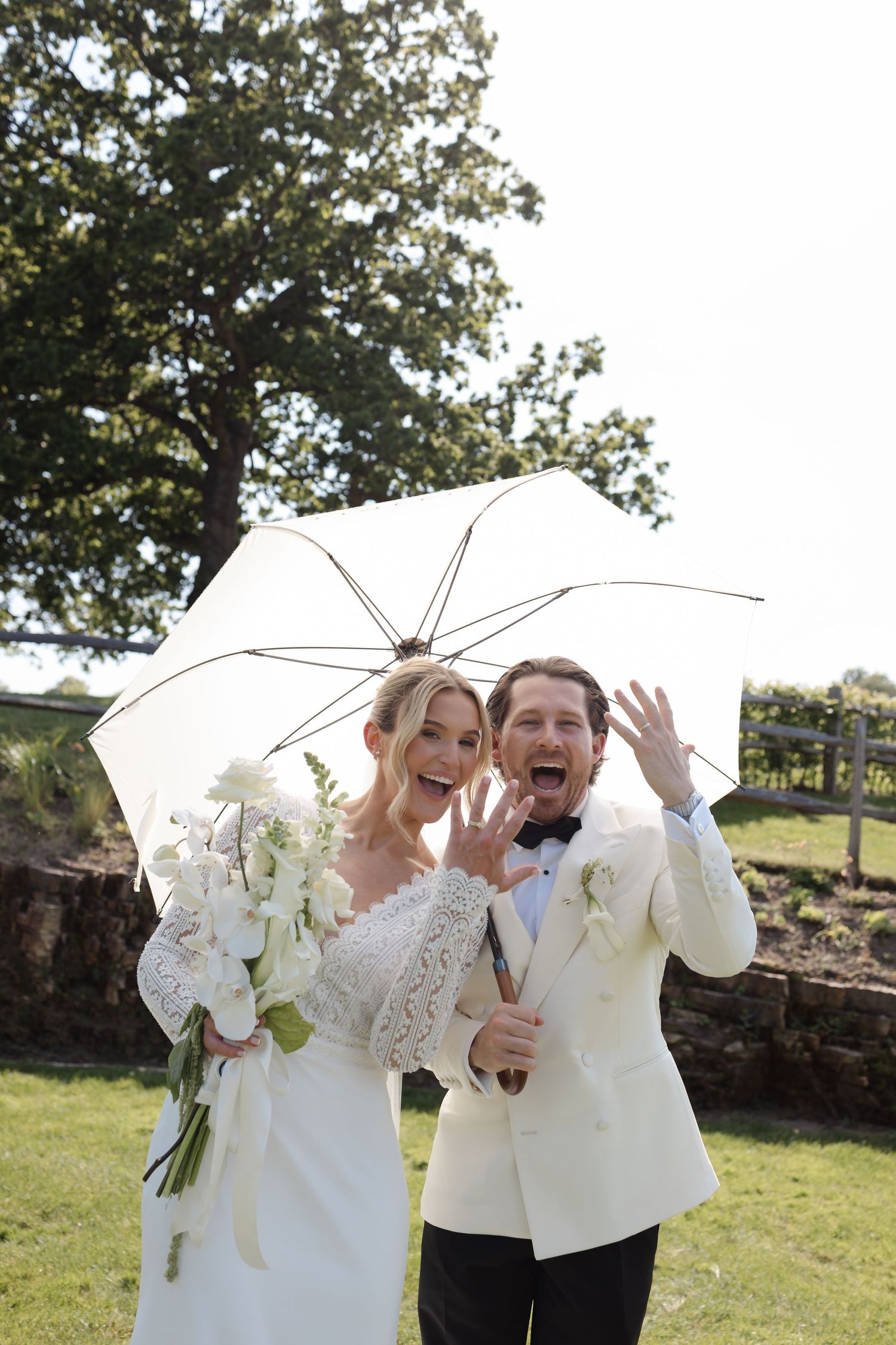 Joyful bride and groom share a white umbrella outdoors, showing off wedding rings with bouquet.