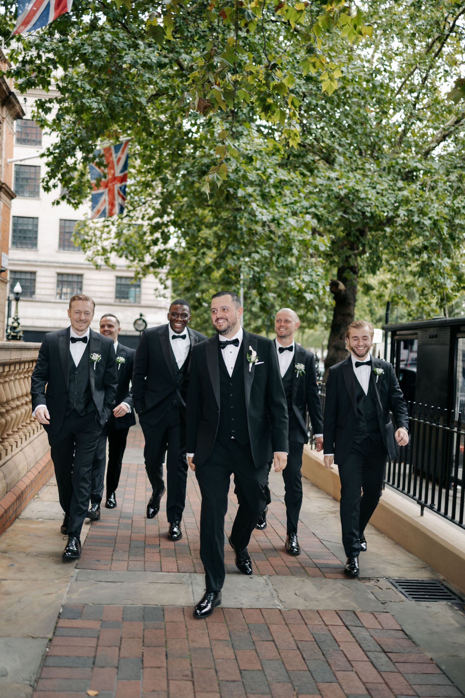 Groomsmen walking together in black tuxedos along brick pathway lined with trees.