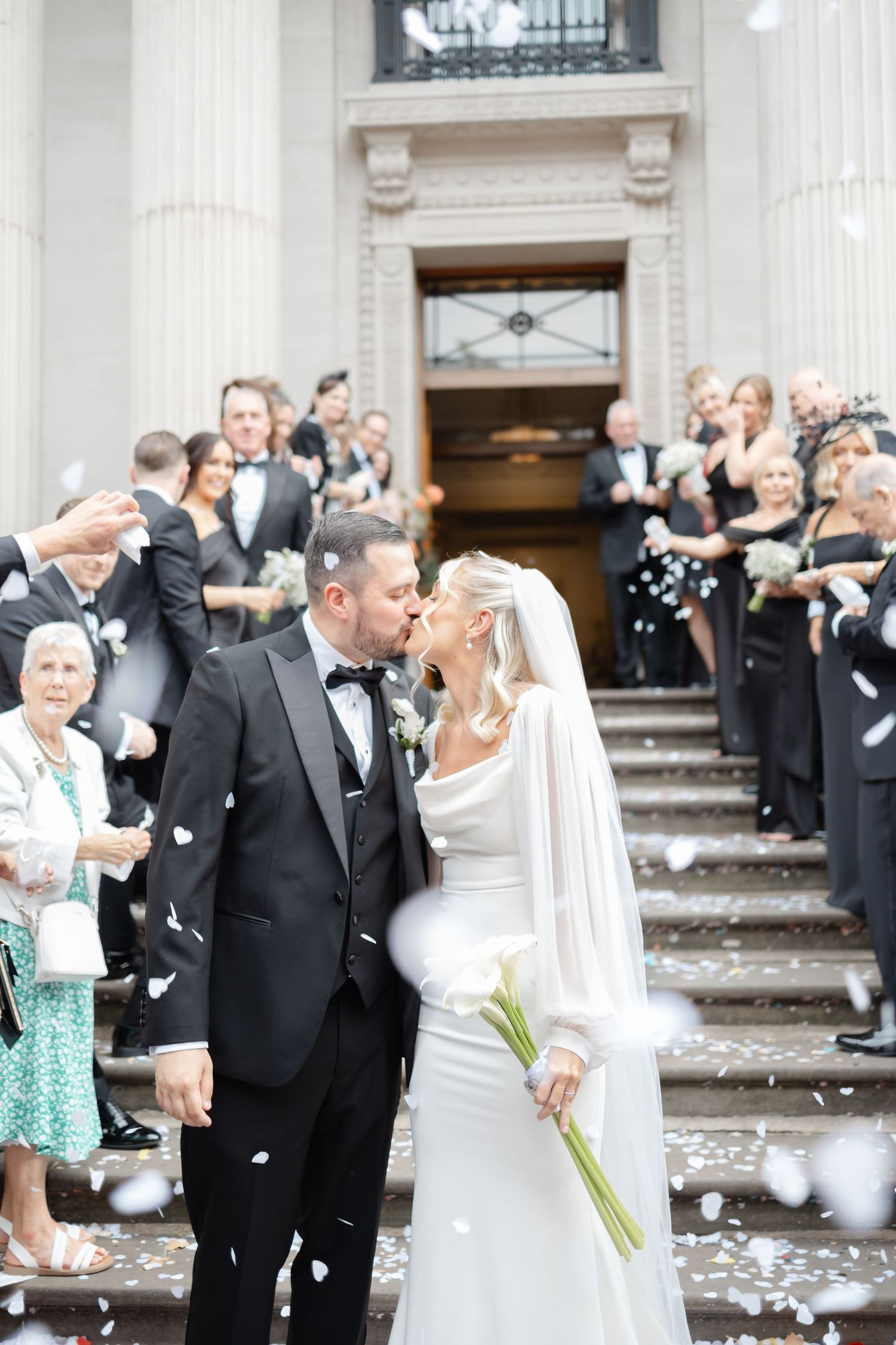 Wedding guests throwing confetti at the newlyweds on the steps of a grand building.