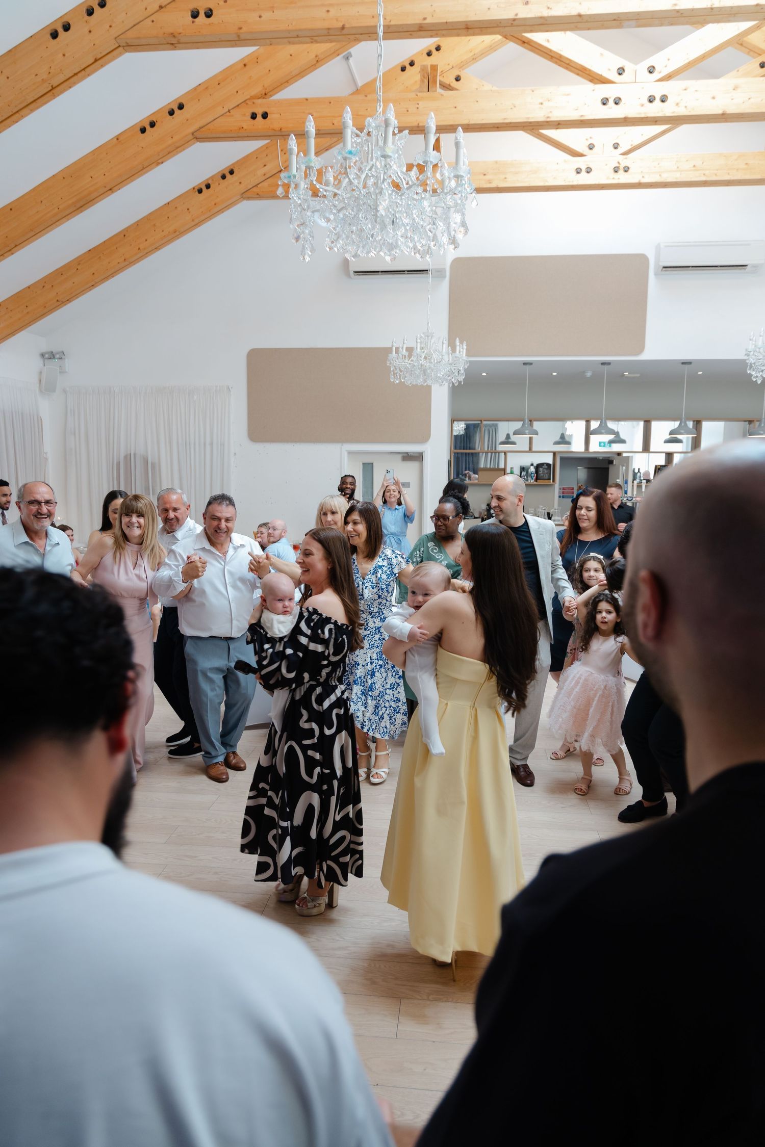 A festive gathering of people dancing in an elegant room with exposed wooden beams and chandeliers.