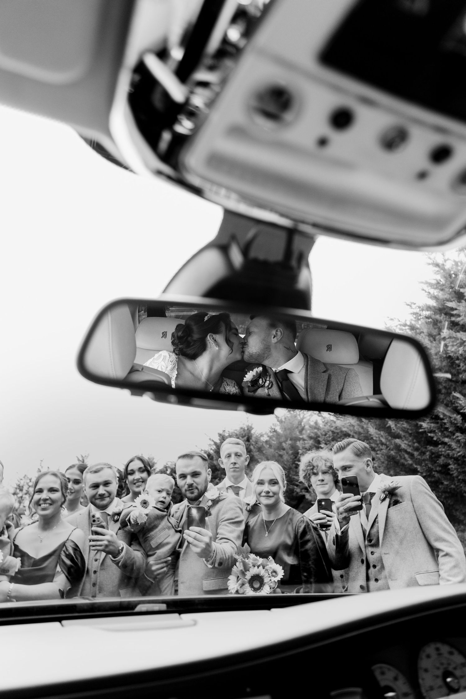 Artistic black and white wedding photo captured through car mirror with wedding party below.