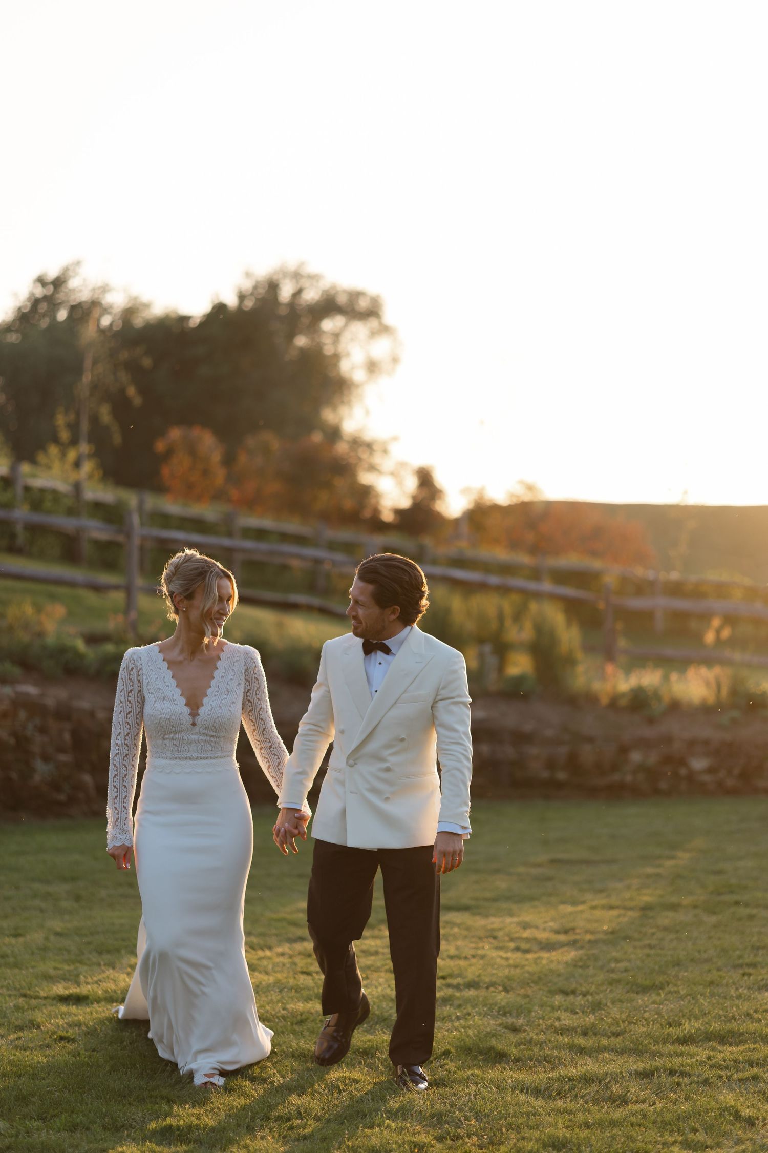 Bride and groom holding hands walking on green lawn at golden hour sunset, she in lace gown, he in white tuxedo jacket.