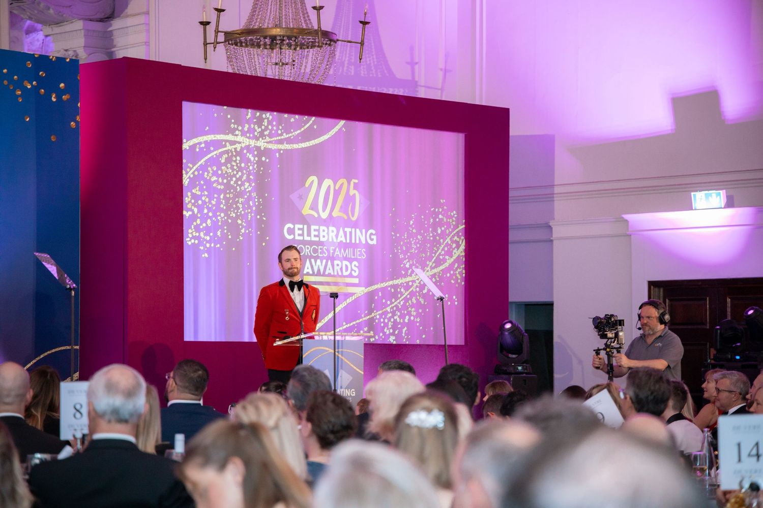 Celebration event with large crowd watching presentation screens in purple-lit ballroom.
