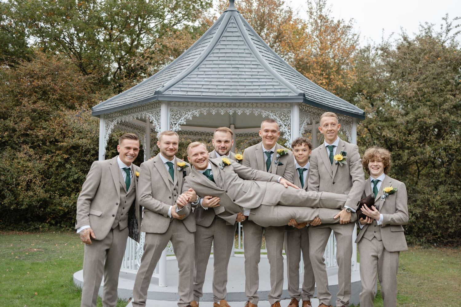 Wedding party in light gray suits posing playfully while holding someone horizontally in front of a garden gazebo.