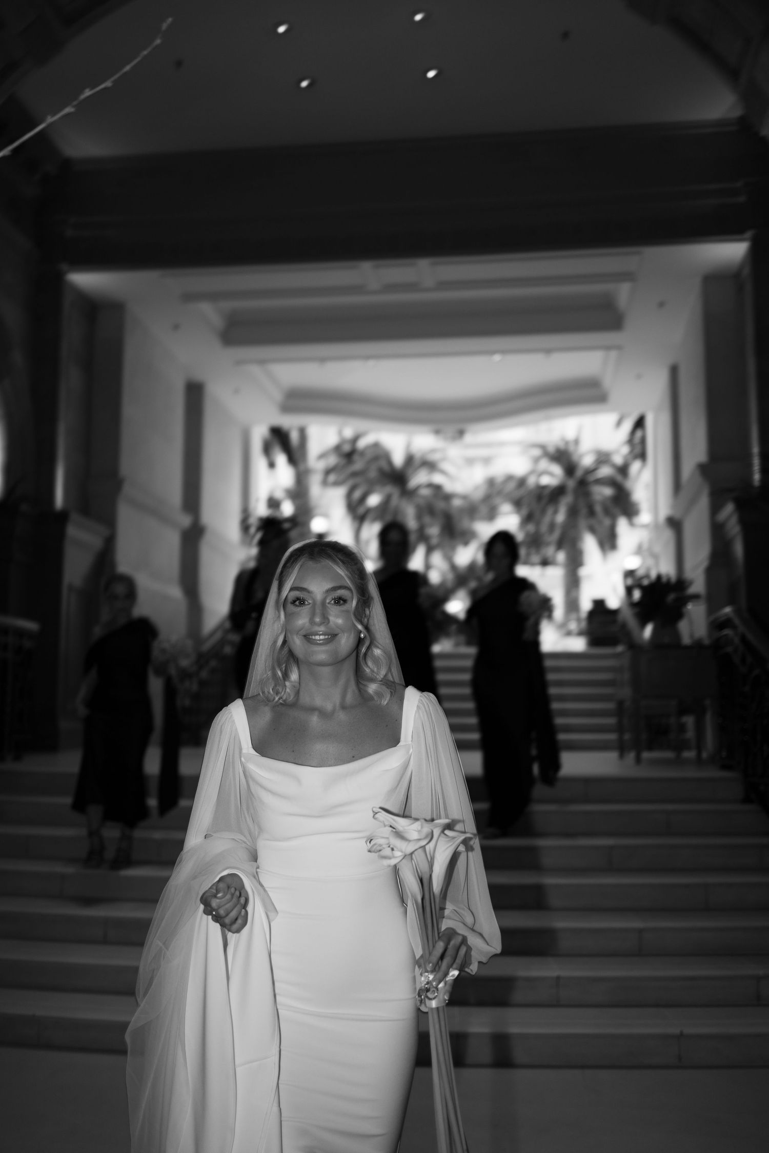 Black and white photo of bride in elegant white dress with long sleeves standing in grand entrance hall.