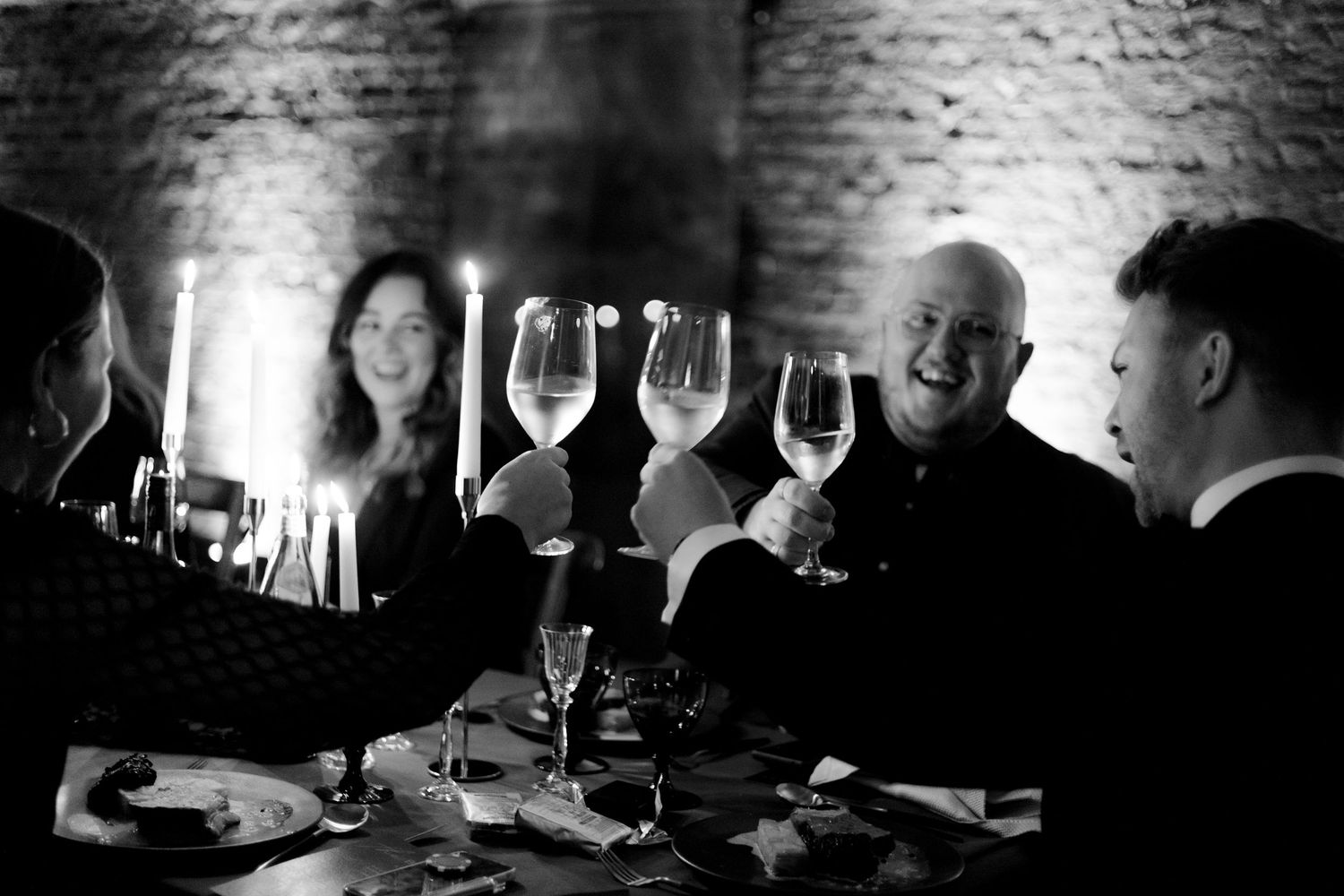 Guests enjoying a candlelit dinner party with wine glasses in a rustic brick-walled venue.