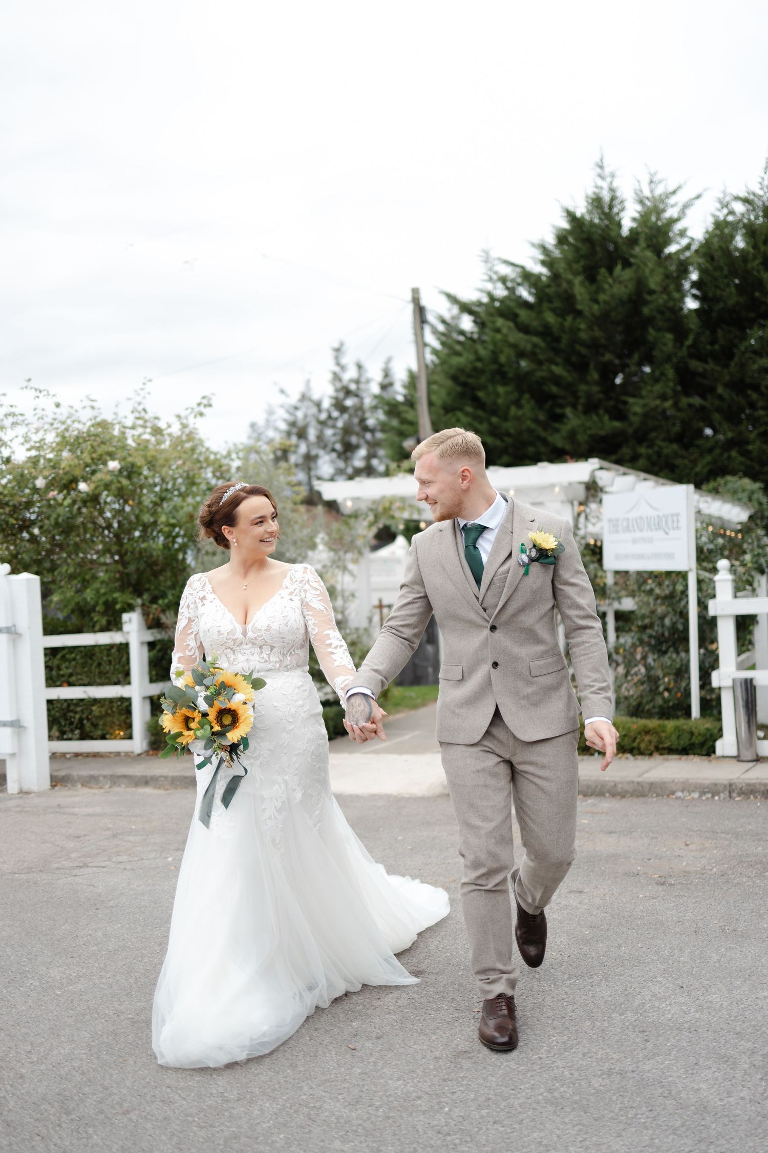 Newlyweds in formal attire skip joyfully down gravel path at wedding venue.