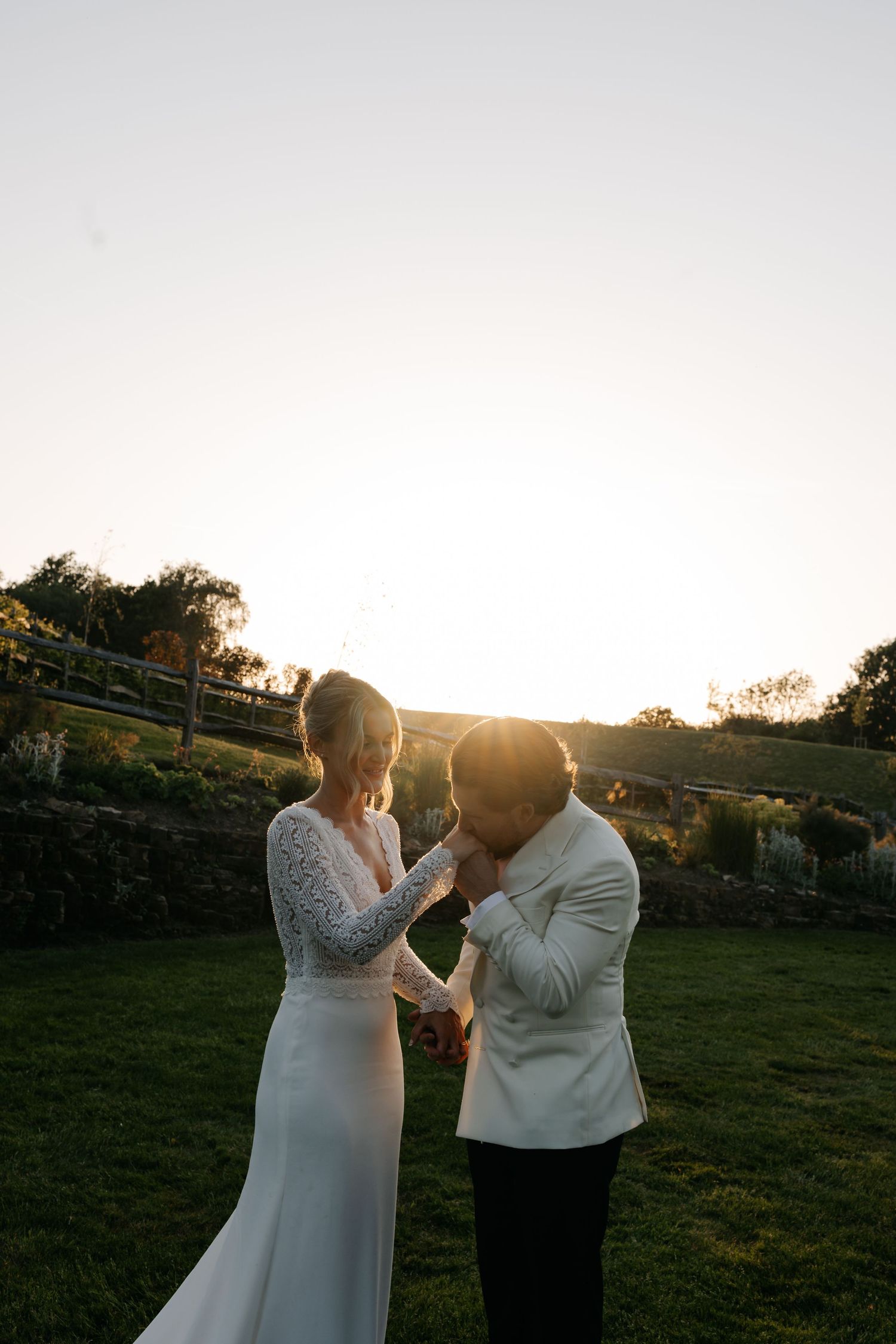 Groom in white jacket kisses bride's hand at golden hour sunset on lush green lawn at outdoor wedding.