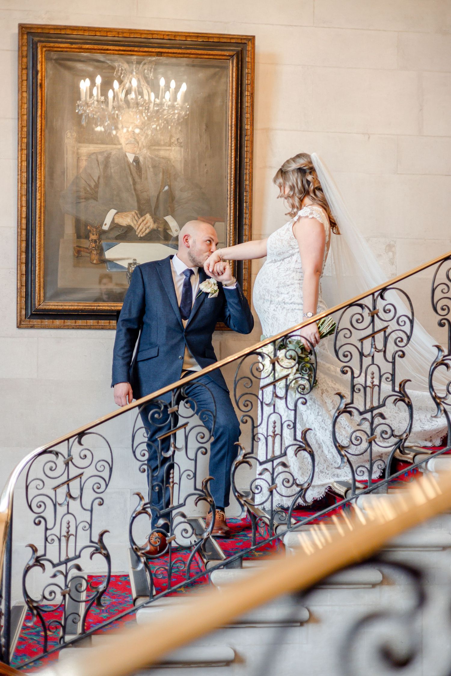 Couple poses playfully on an elegant staircase with ornate iron railings and framed artwork.