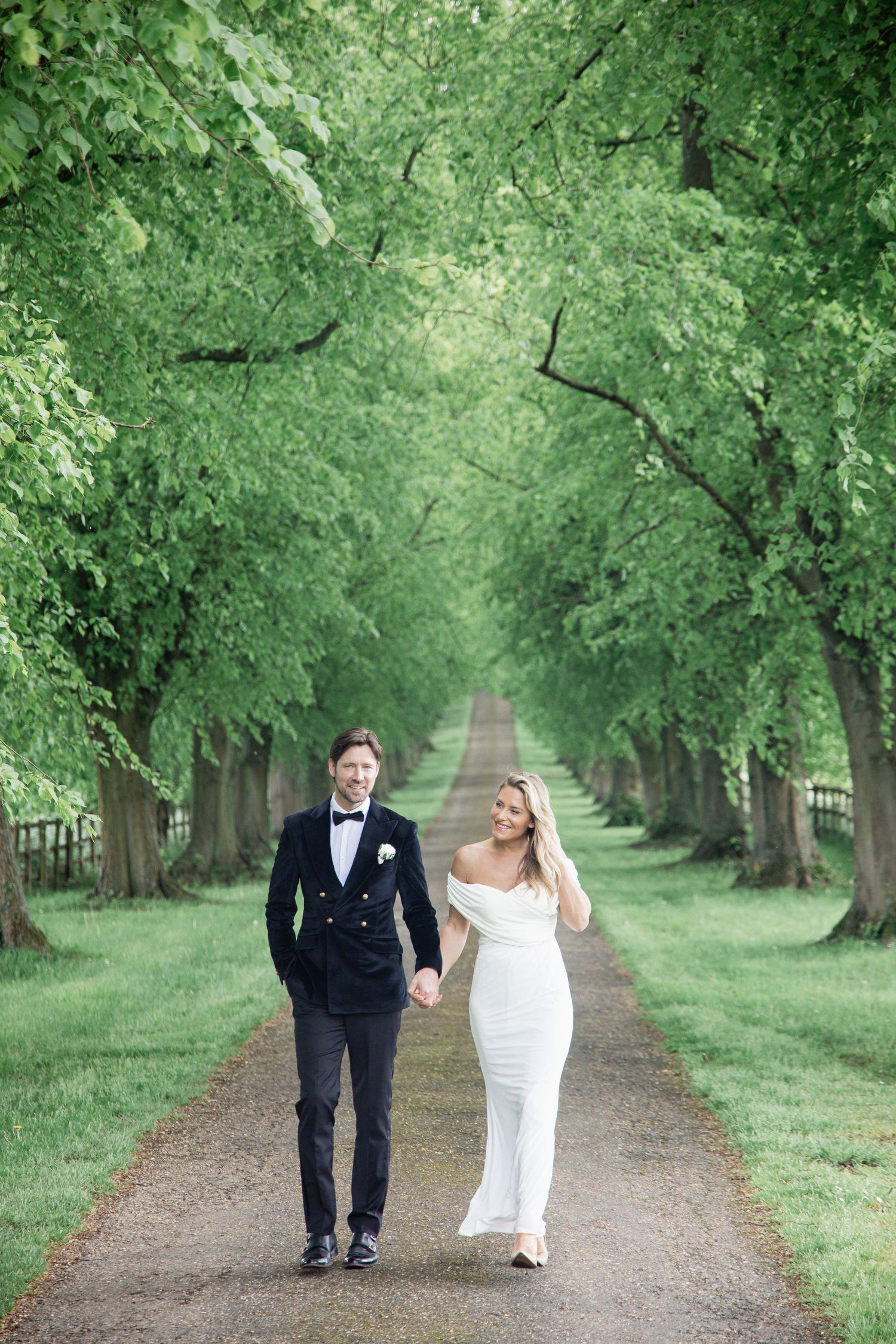 A couple in formal attire walks down a tree-lined path surrounded by lush green foliage.