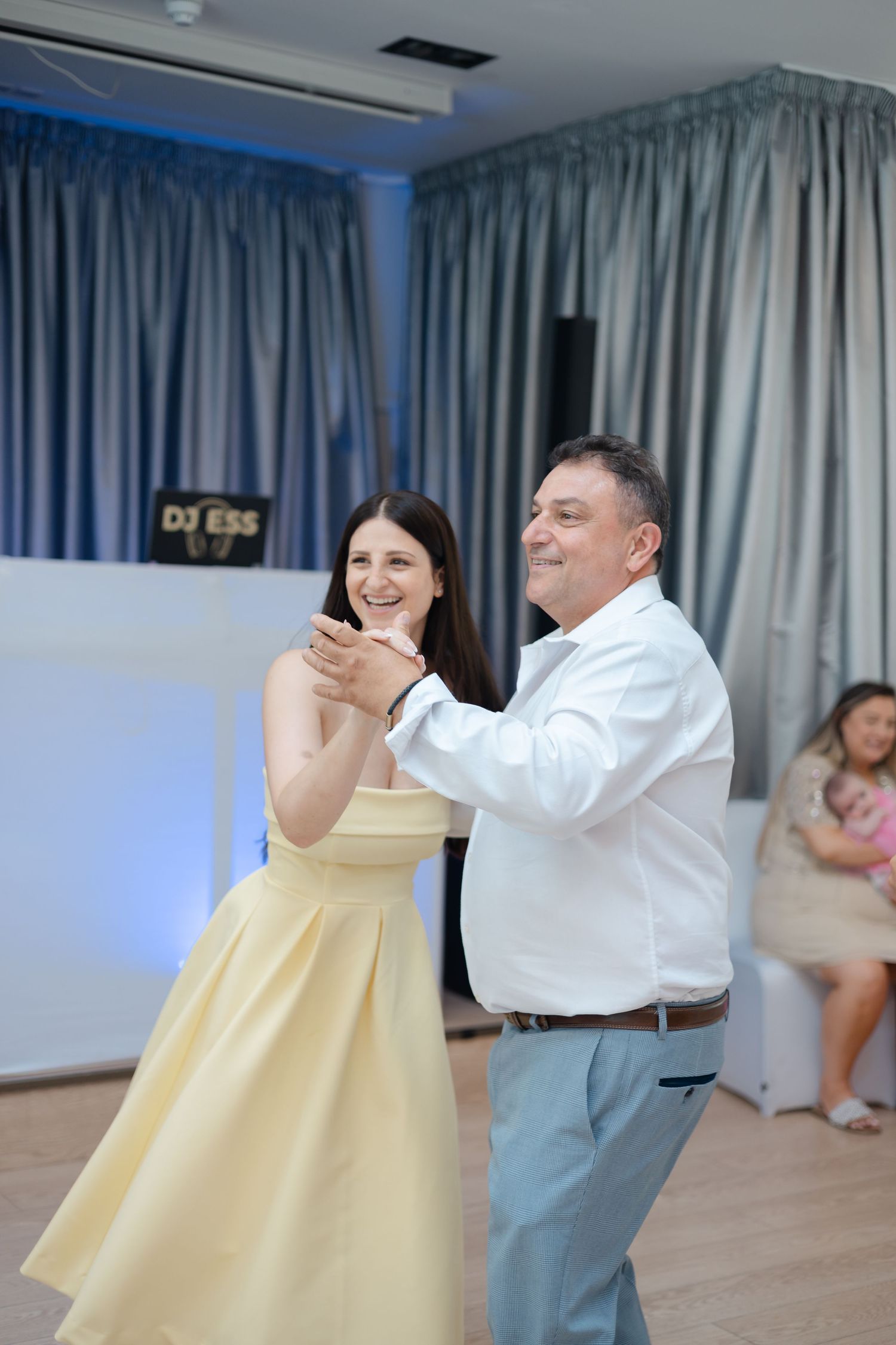 A couple dances together at an event, with the woman wearing a pale yellow dress and the man in a white shirt.