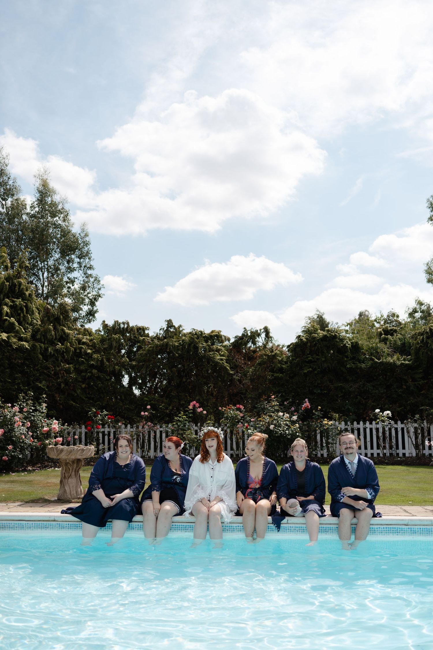 Wedding party sitting at the edge of a swimming pool with their feet in the water on a sunny day.