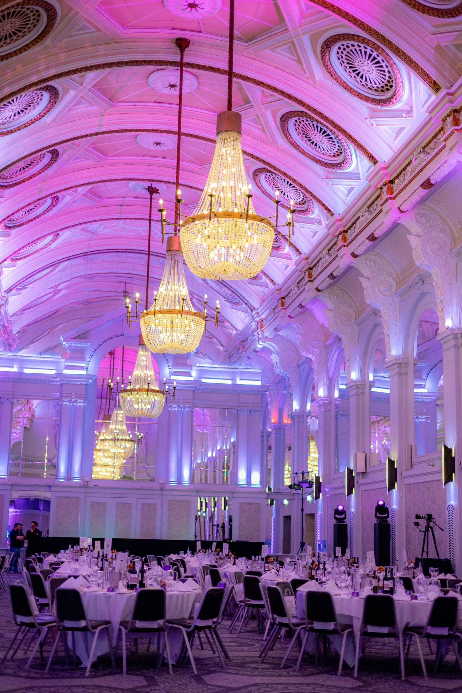 Elegant ballroom with ornate chandeliers and pink uplighting illuminating white columns and table settings.
