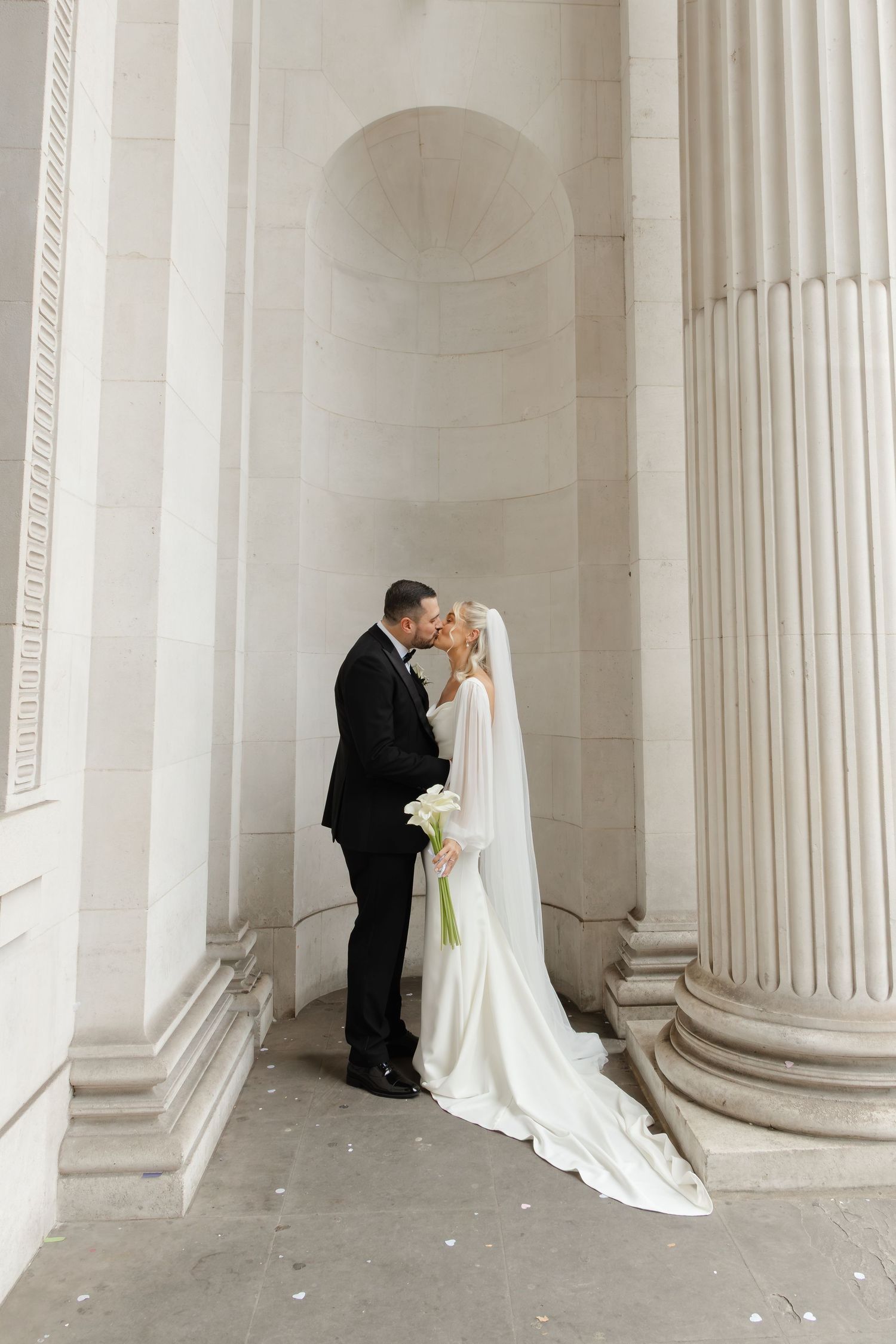 A romantic wedding photo of a couple sharing an intimate moment between elegant white columns.