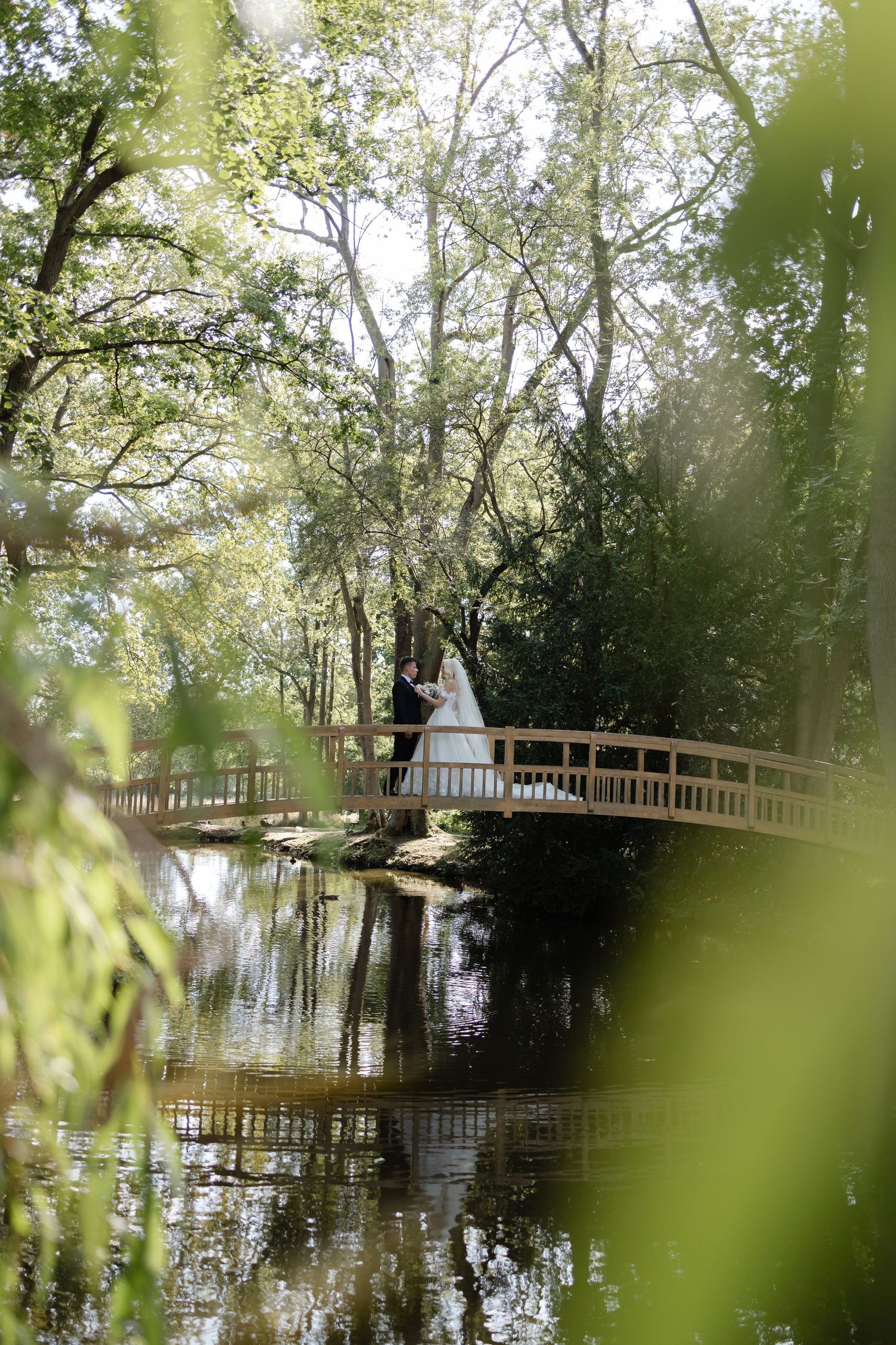 Romantic wedding portrait on wooden bridge over reflective pond surrounded by lush greenery.