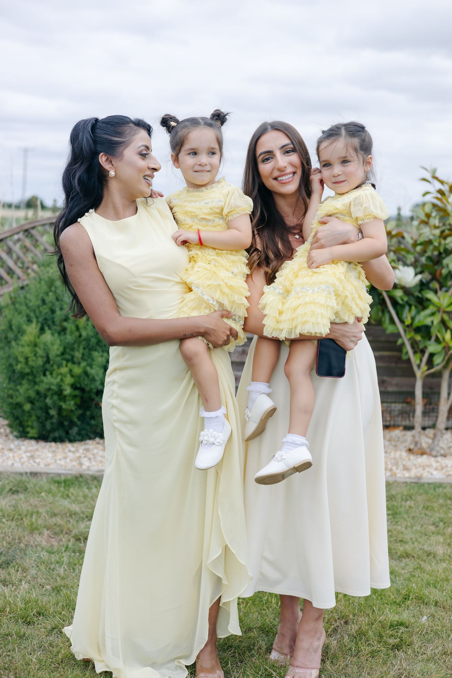 Women in pale yellow dresses hold small children in matching outfits on a sunny day.