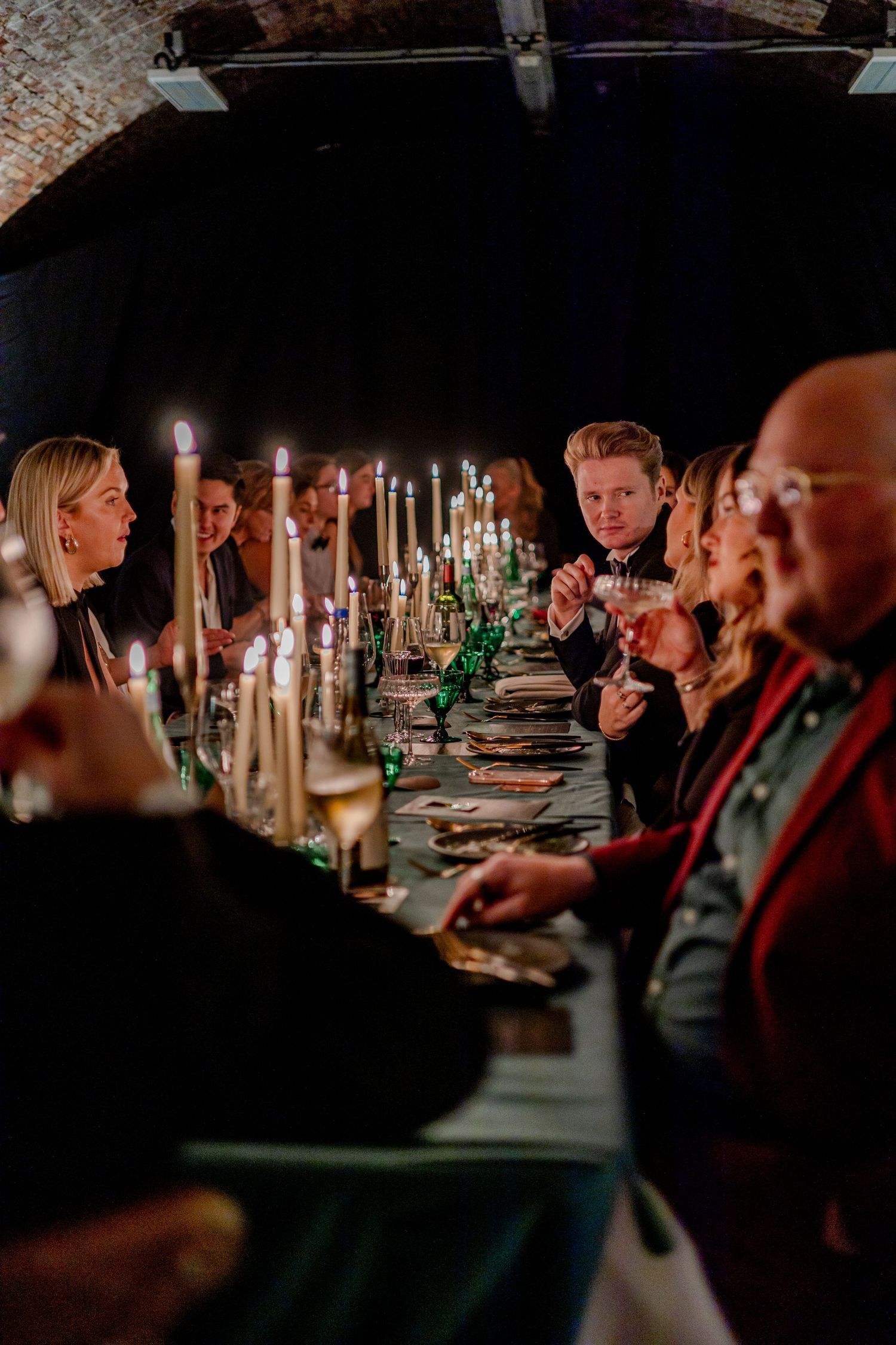 Guests seated at an elegant candlelit dinner table in a rustic brick-walled venue.