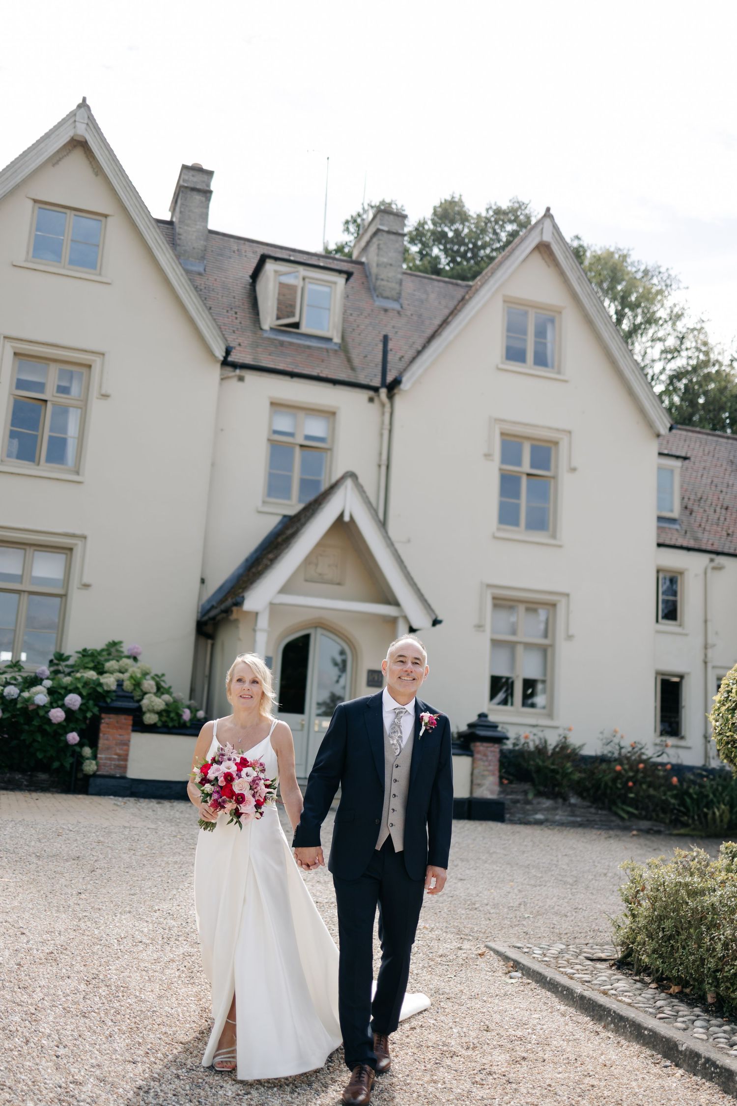 Wedding couple walks along gravel path outside elegant cream-colored country house with multiple gabled windows.