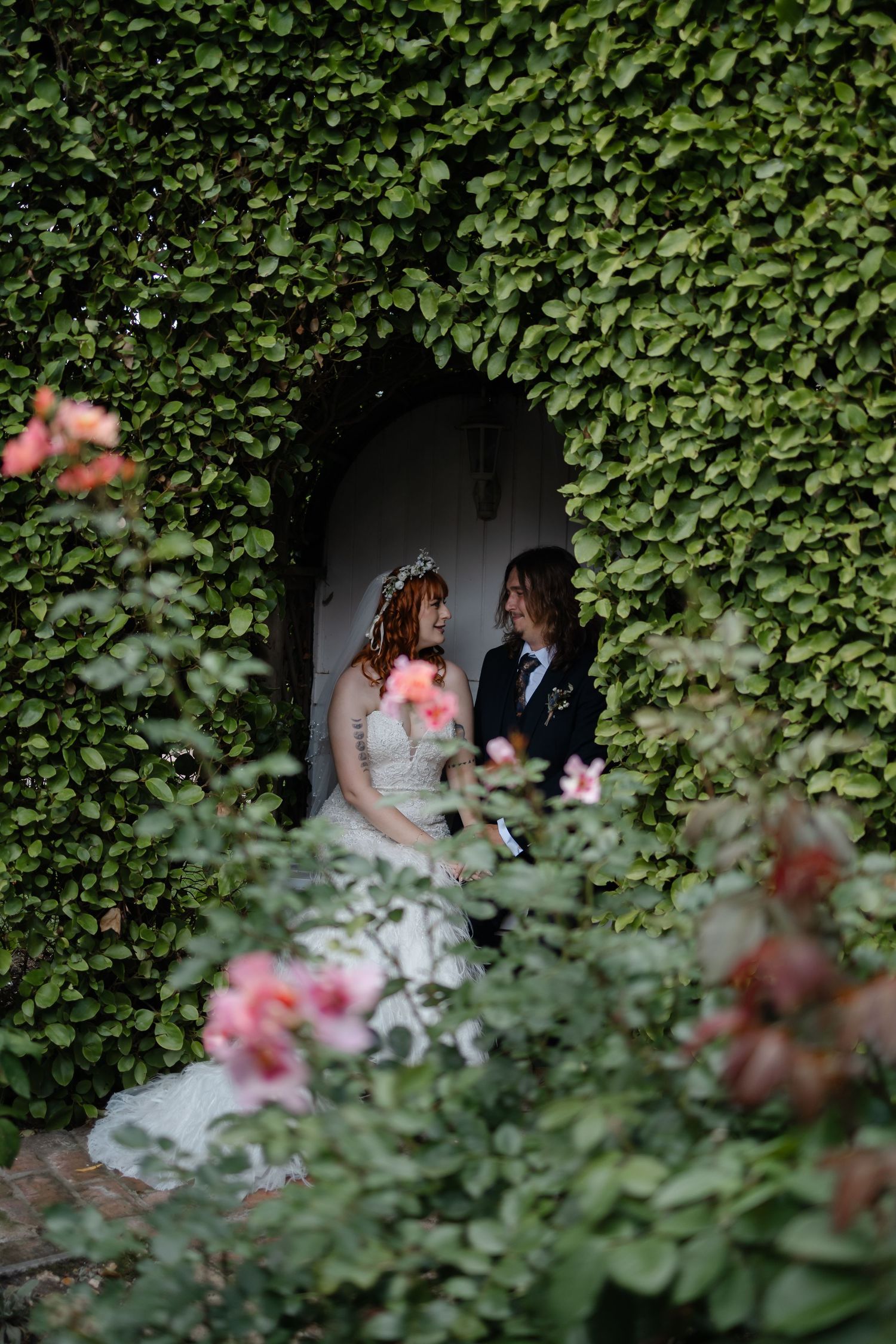 A romantic wedding couple framed by ivy and pink flowers in a garden doorway.