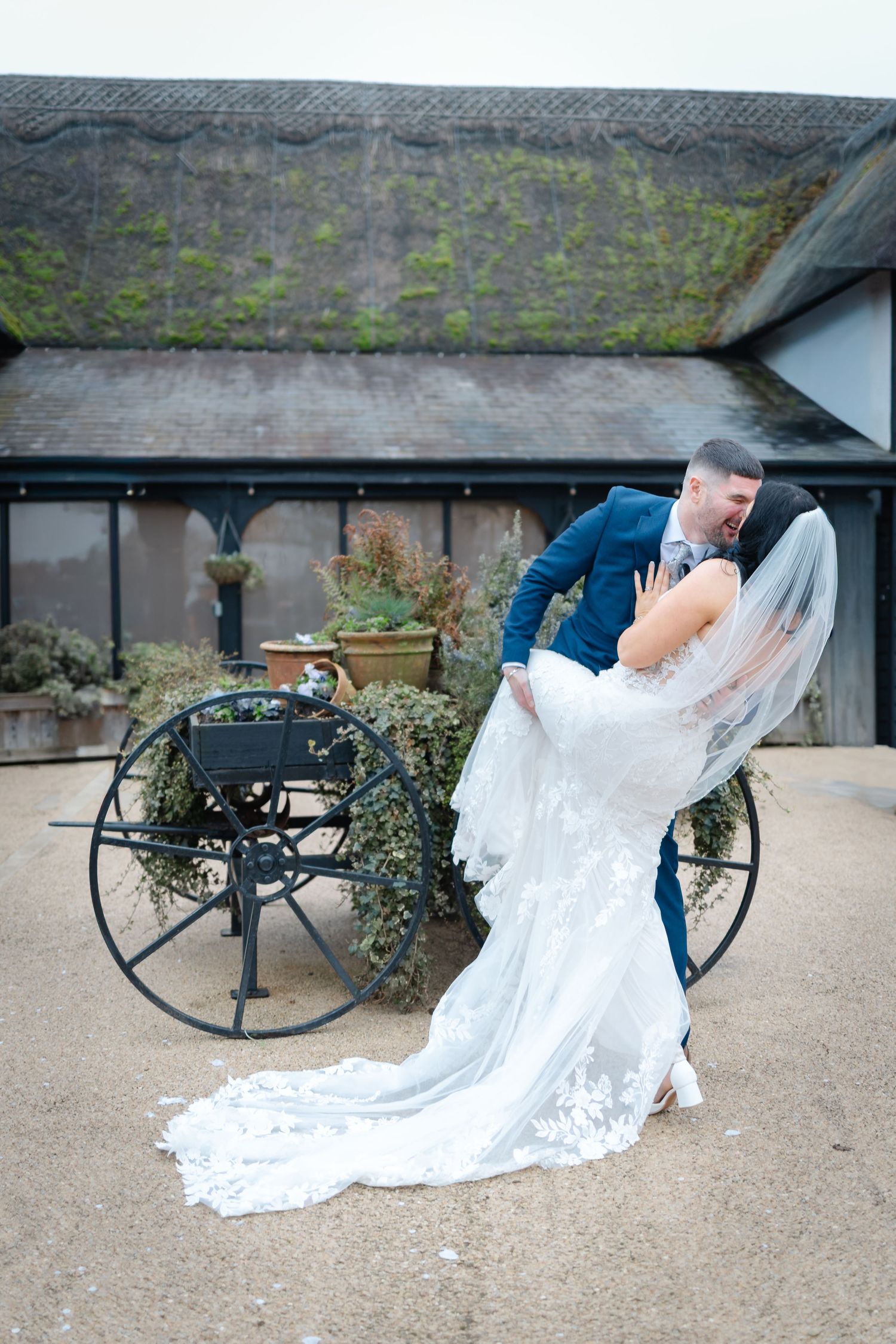 Wedding couple poses near vintage wagon wheels at a rustic outdoor venue.