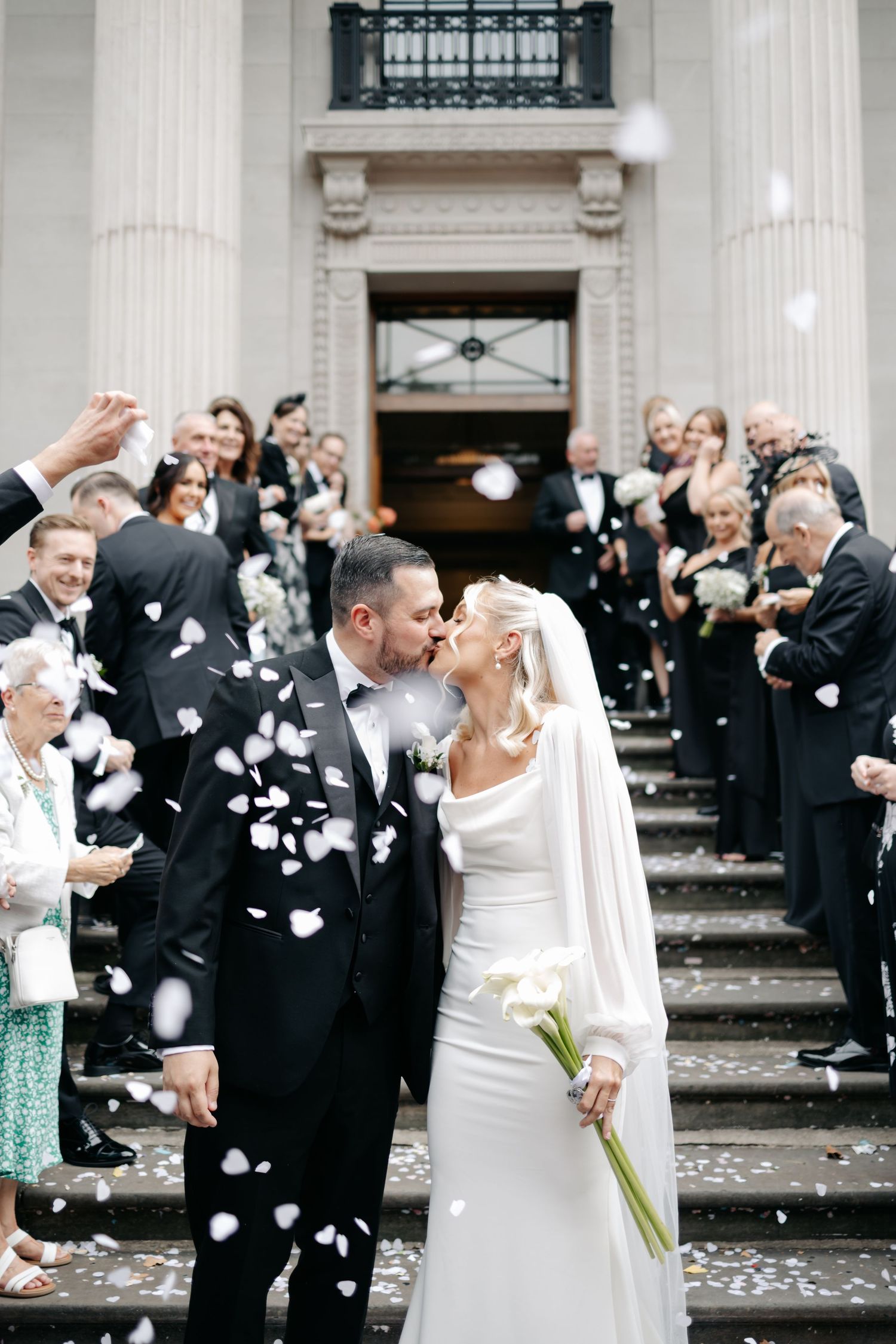 Wedding guests toss white flower petals during an outdoor celebration on grand stone steps.