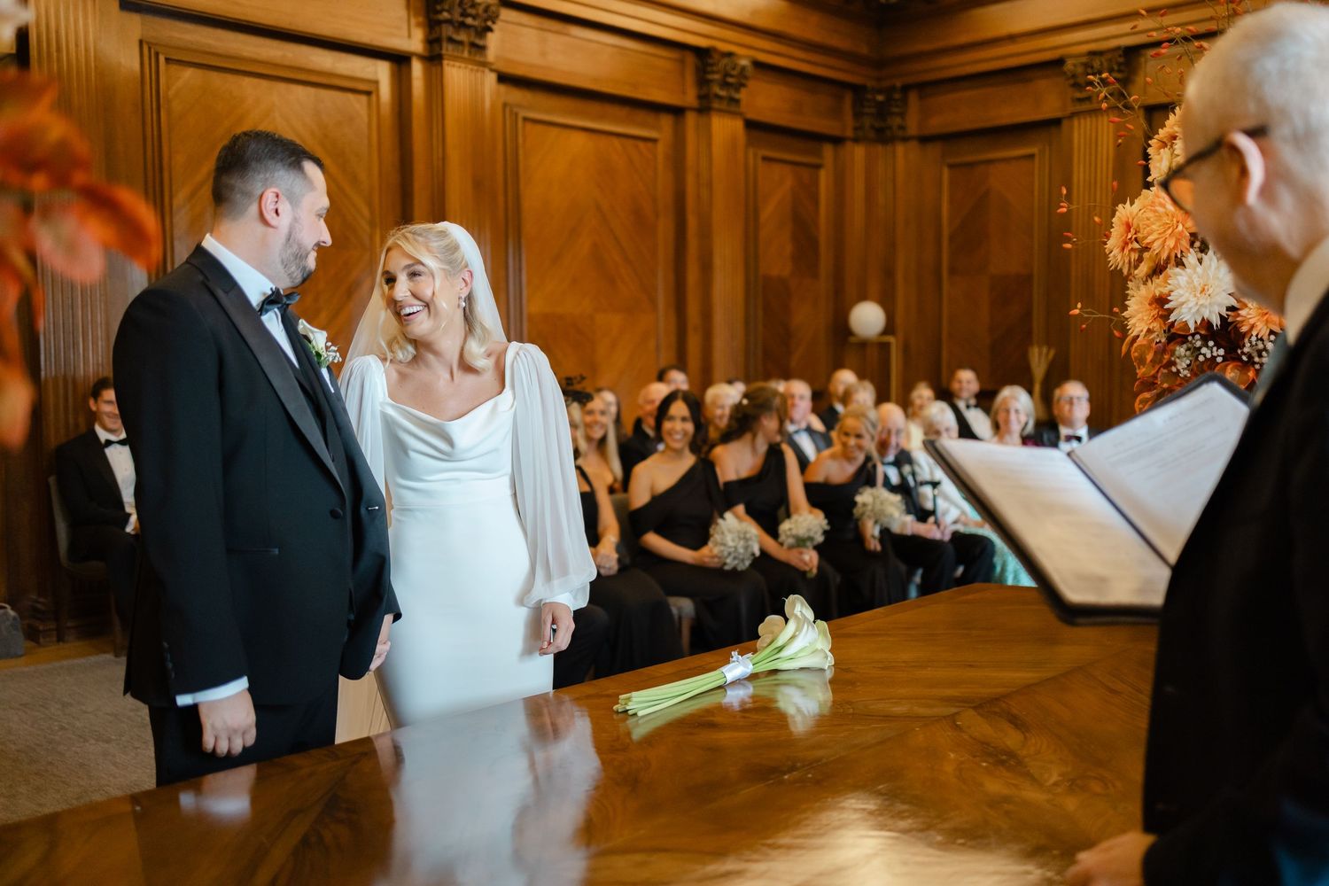 Wedding ceremony in wood-paneled room with guests watching couple exchange vows.