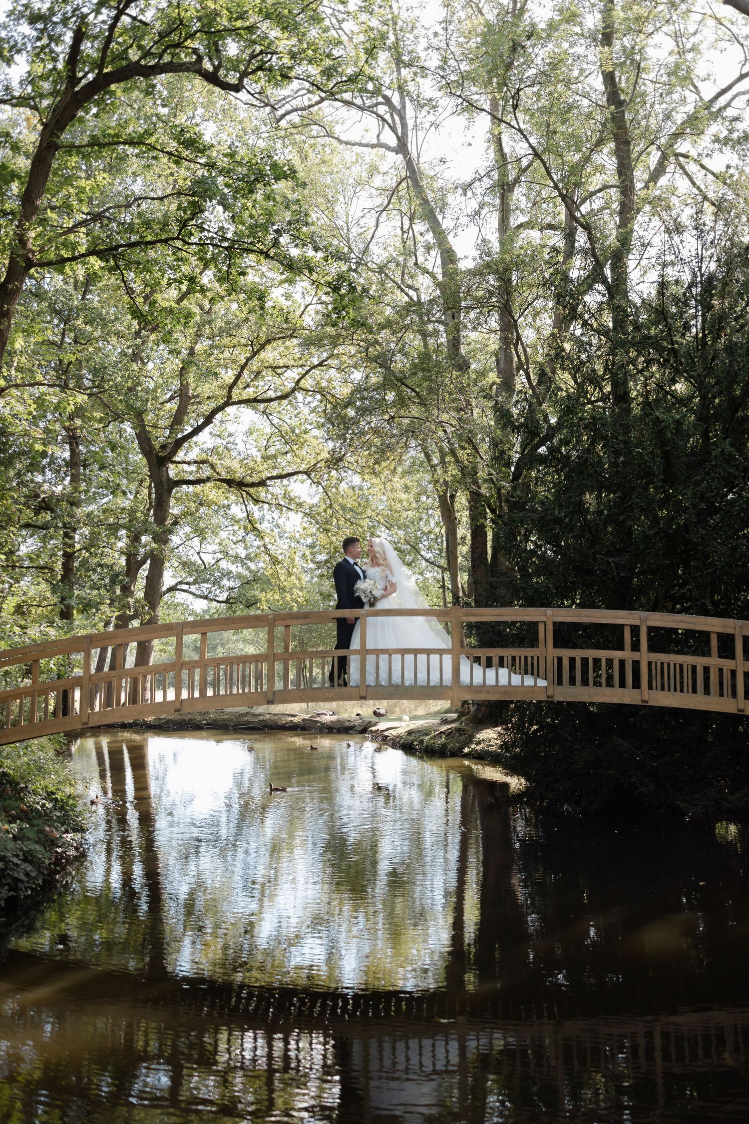 A couple shares a romantic moment on a wooden bridge over reflective water surrounded by trees.