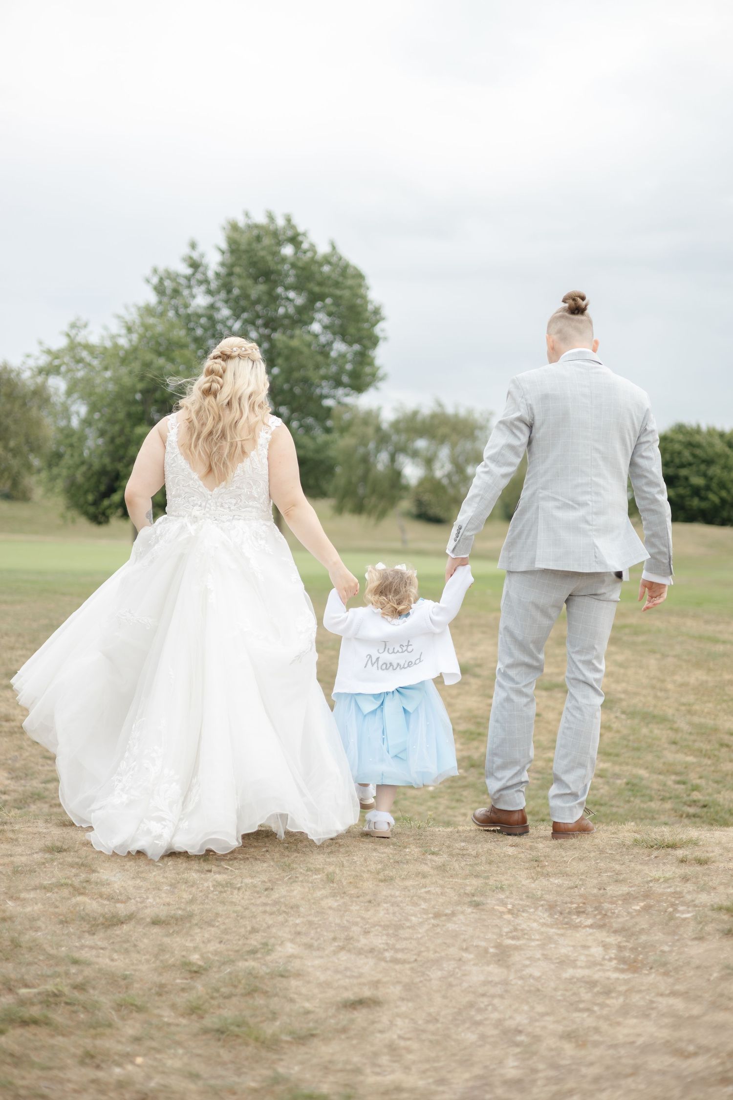 Wedding party walks through a grassy field holding hands in a line against a cloudy sky.