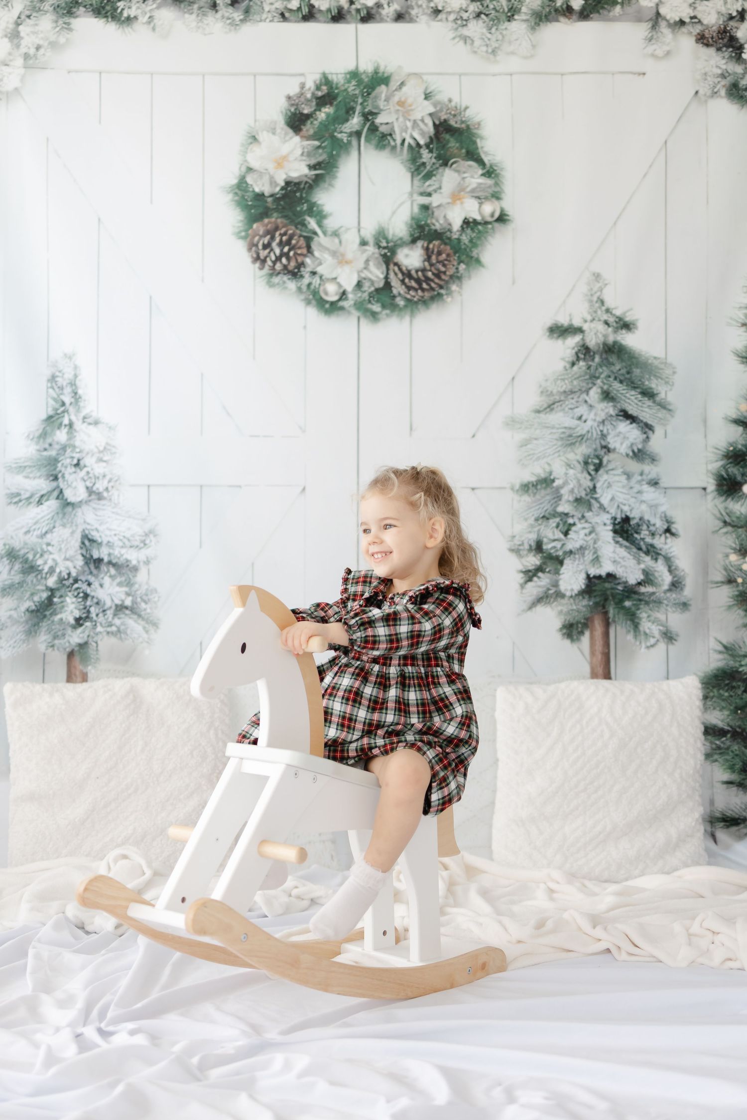 Child rides wooden rocking horse against white winter backdrop with wreath and snowy trees.