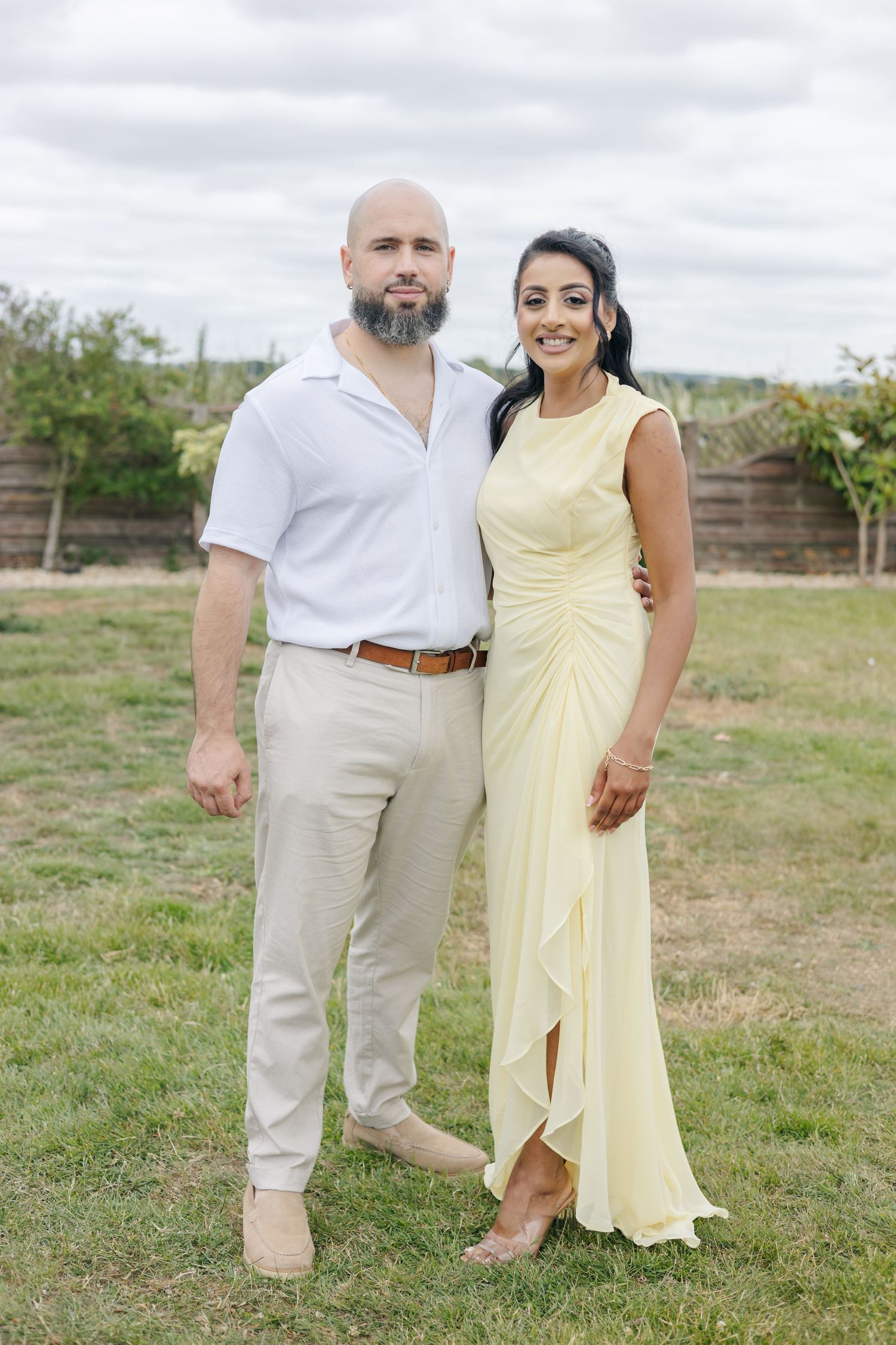 Couple in light summer attire poses together on grassy field under cloudy sky.