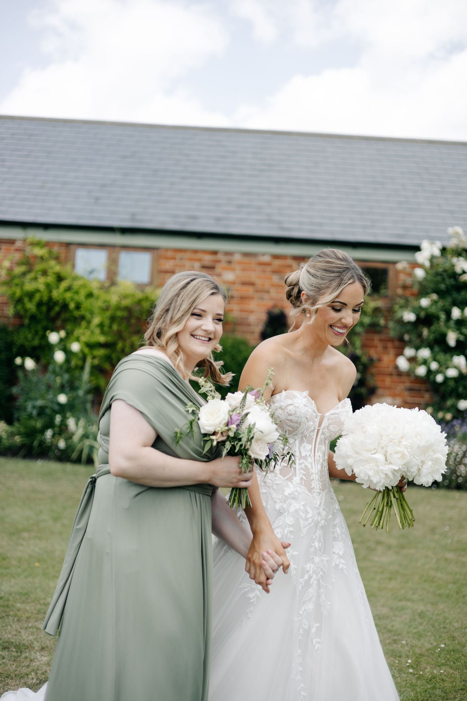 Bride and bridesmaid embrace outdoors against brick building with white flowers in hand.