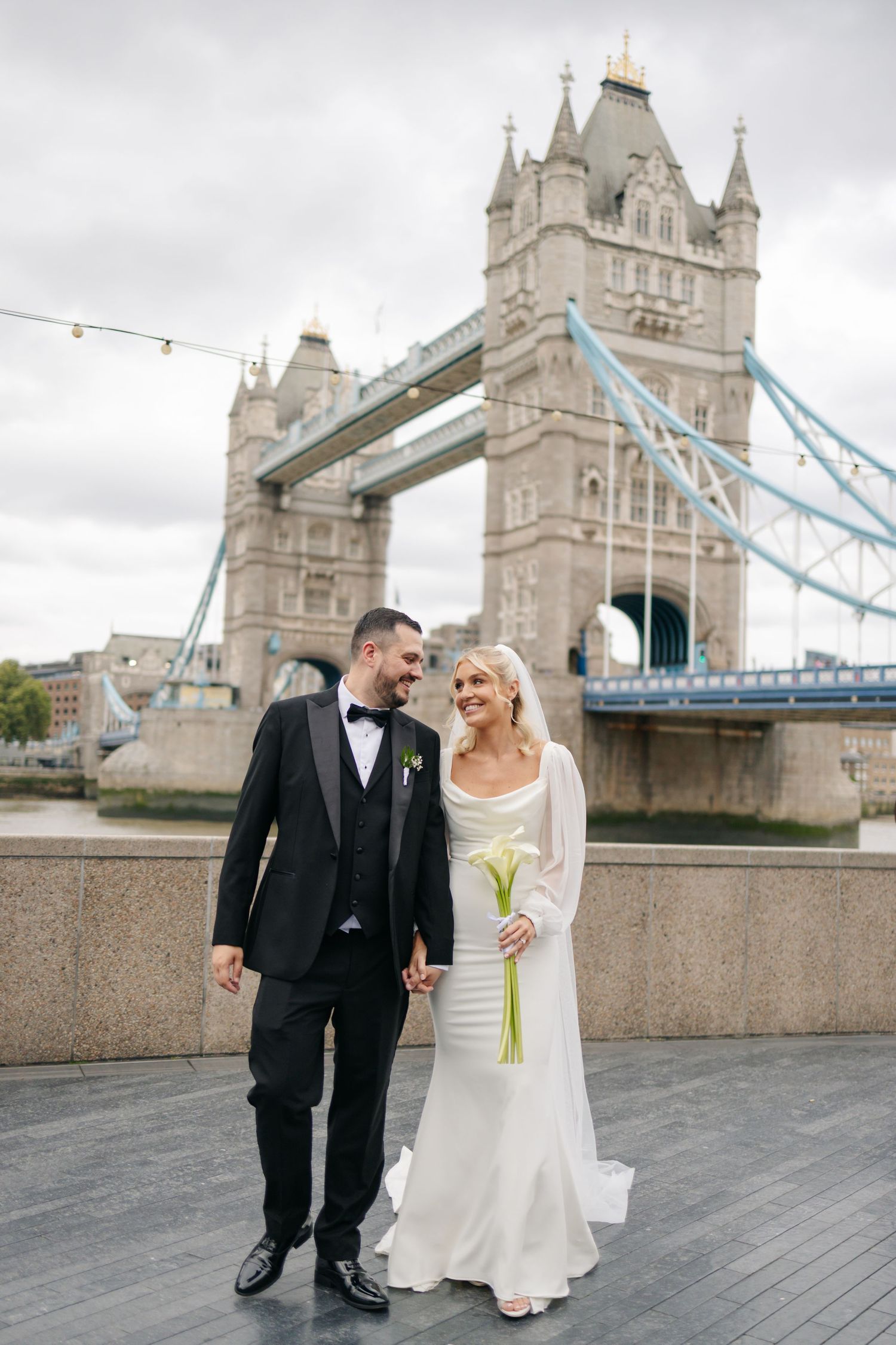 Newlyweds sharing joyful moments along the Thames River with Tower Bridge in background.